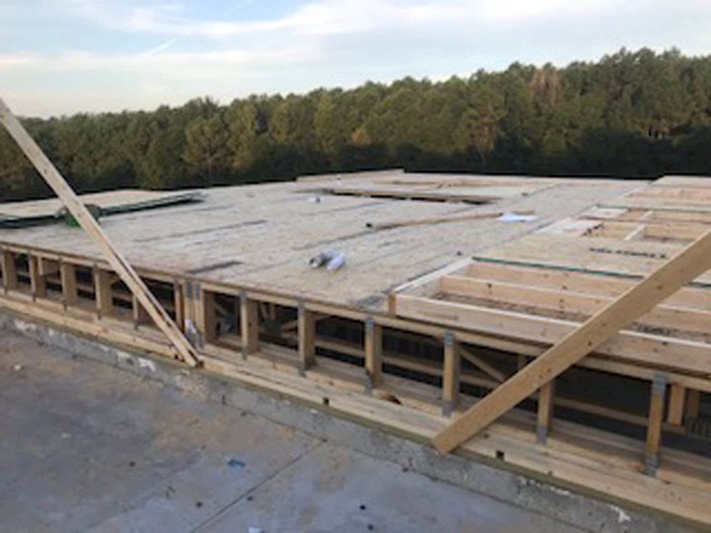 Wood-framed house under construction with exposed beams, open roof, and distant trees against a cloudy sky