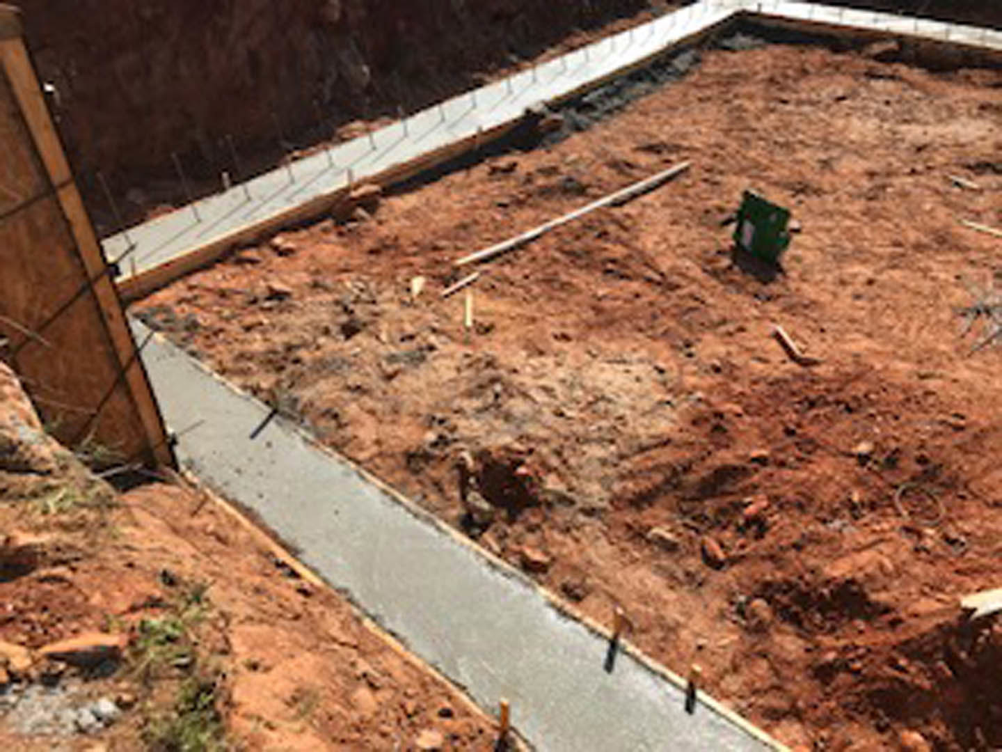 Concrete foundation set in exposed dirt, with nearby green vegetation and wood panel detail visible.