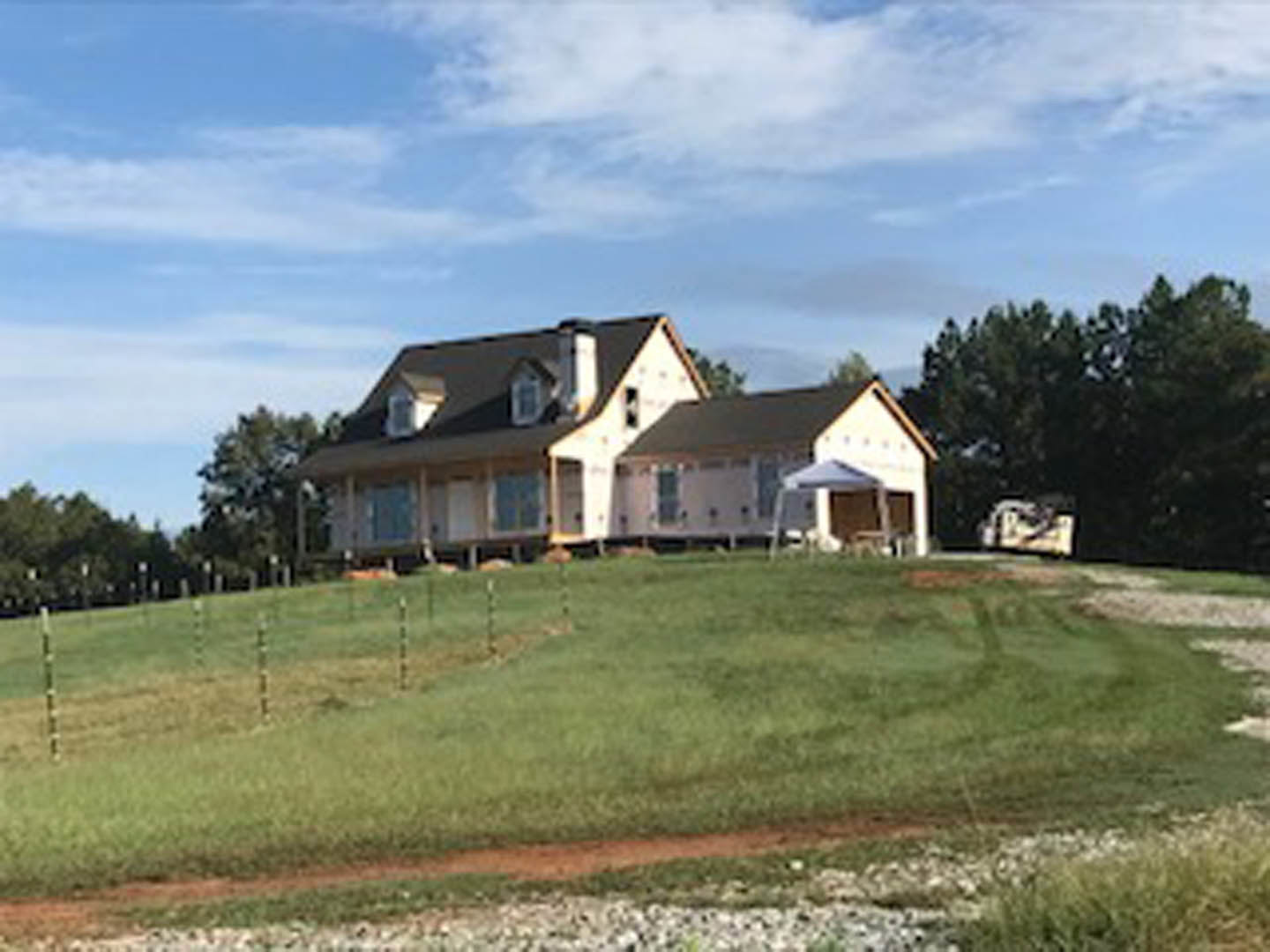 Two-story farmhouse with white siding and brick chimney, set atop a grassy hill, surrounded by trees and a wooden fence under a partly cloudy sky