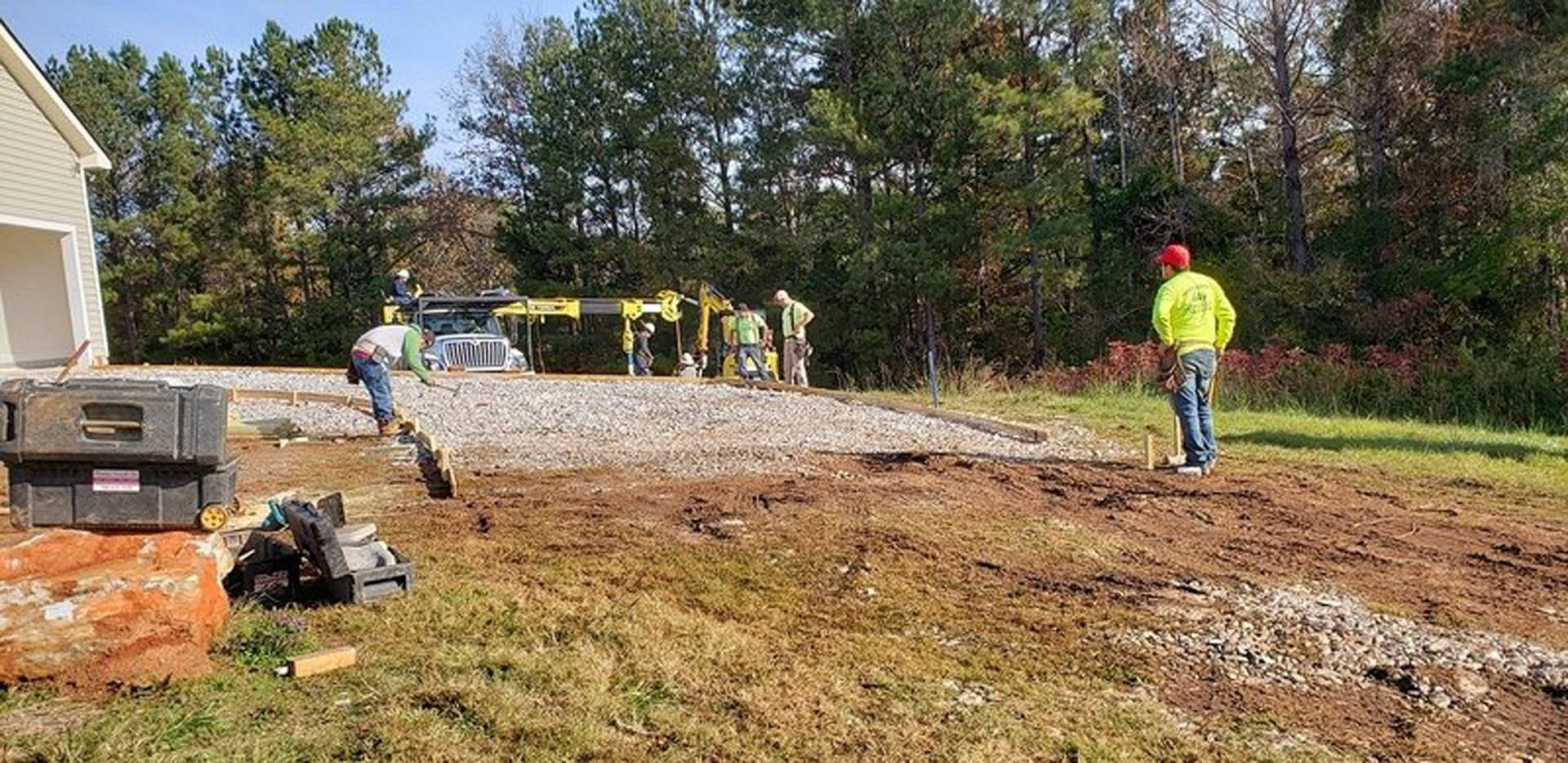 Workers in yellow vests standing on soil near a partially built house with exposed framing, surrounded by grass, trees, and construction materials under a clear sky