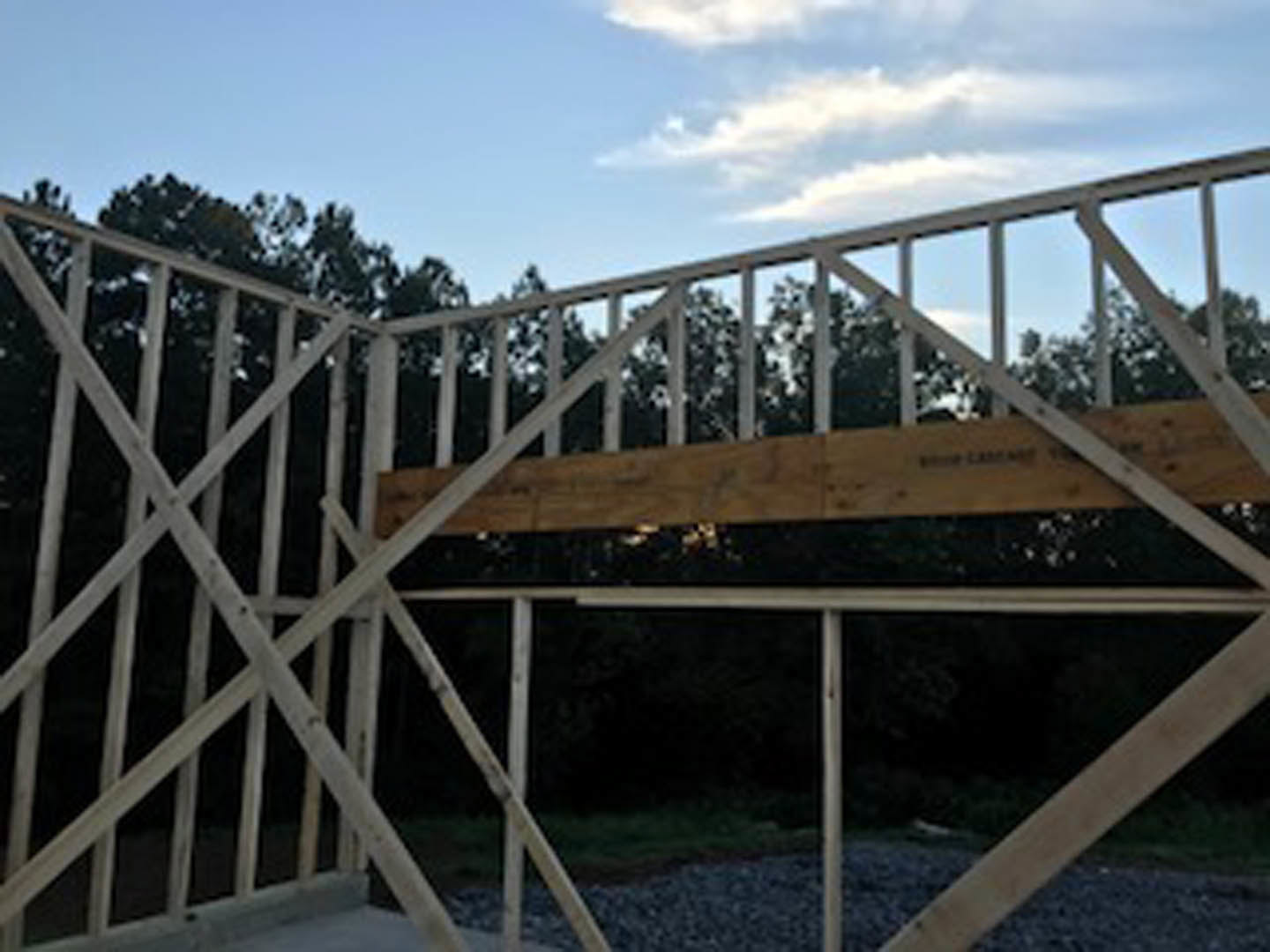 Exposed wooden beam on a deck with white X-shaped railing, blue sky and trees in background