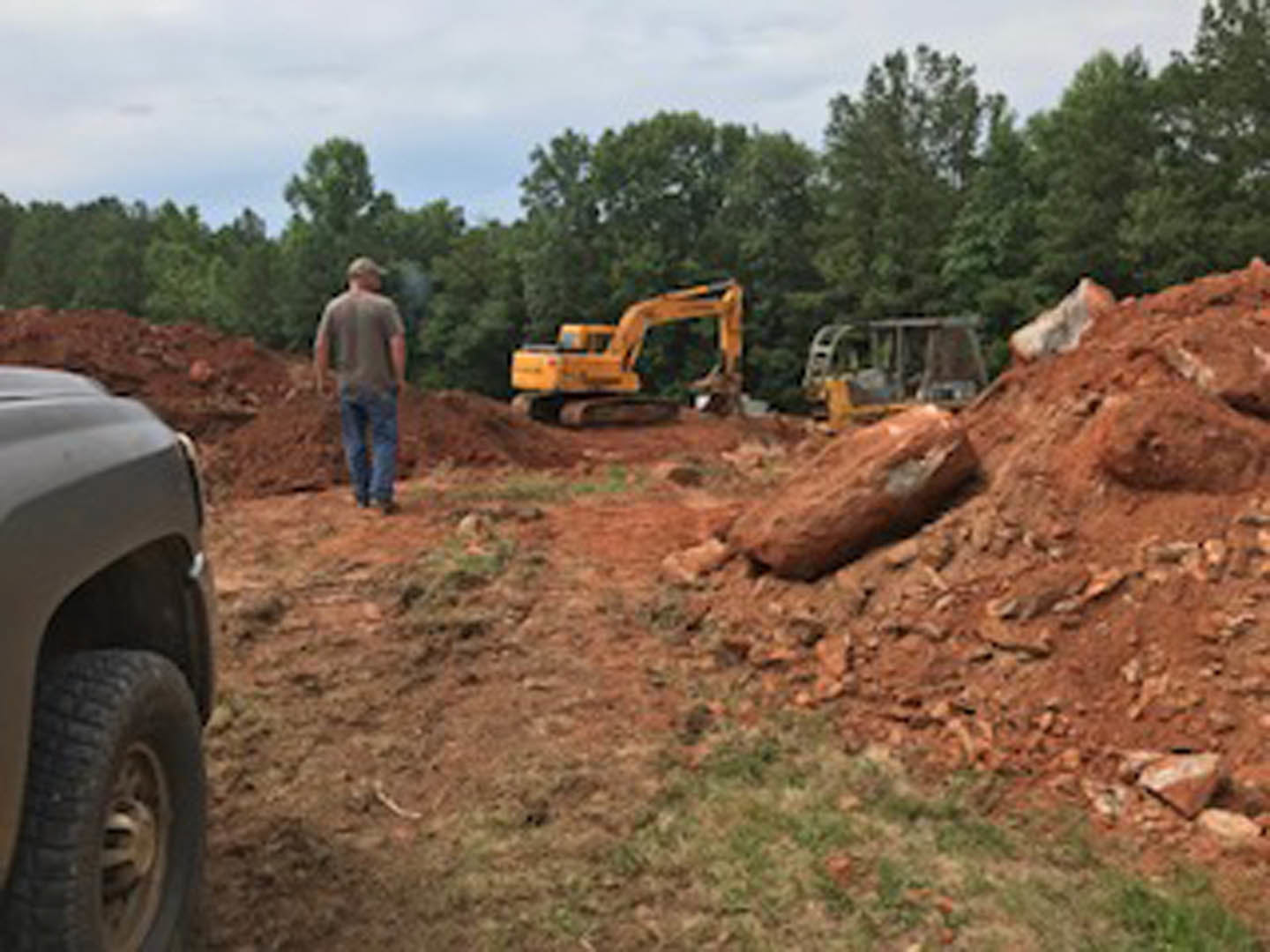 Man standing on dirt lot with yellow bulldozer and tractor, surrounded by soil, trees, and open sky
