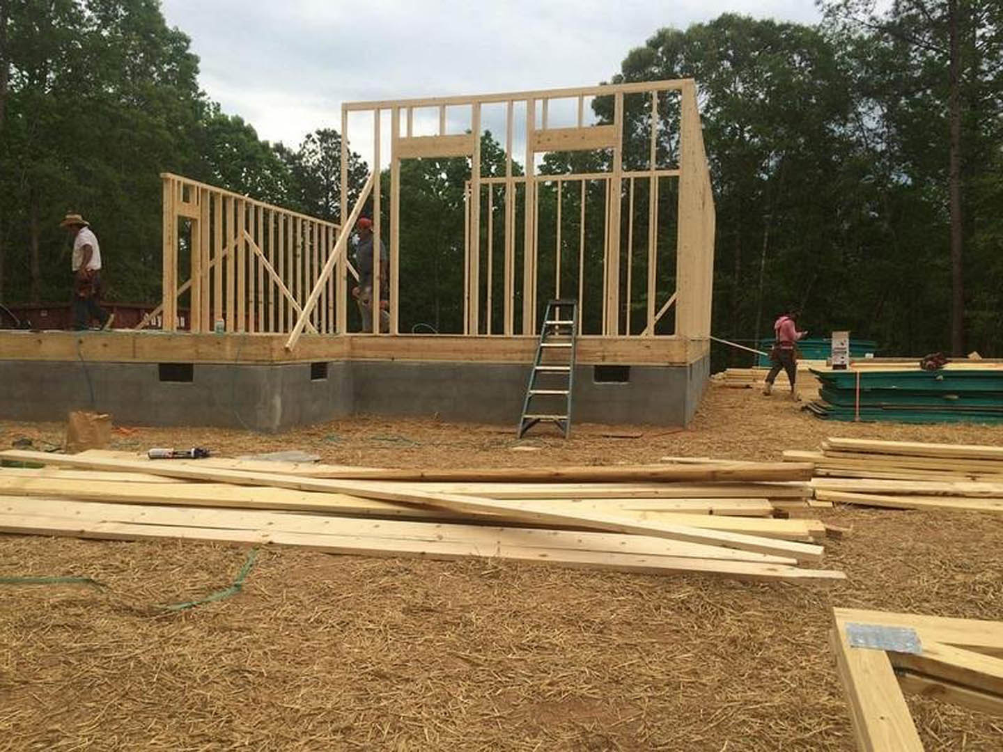 Partially built brick house with exposed framing, metal ladder leaning against wall, two men working near construction materials, green and yellow bench in foreground, trees and