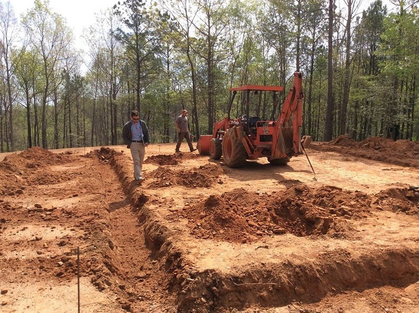Man standing beside yellow tractor on dirt lot with scattered soil, trees in background, clear sky overhead, construction equipment visible.