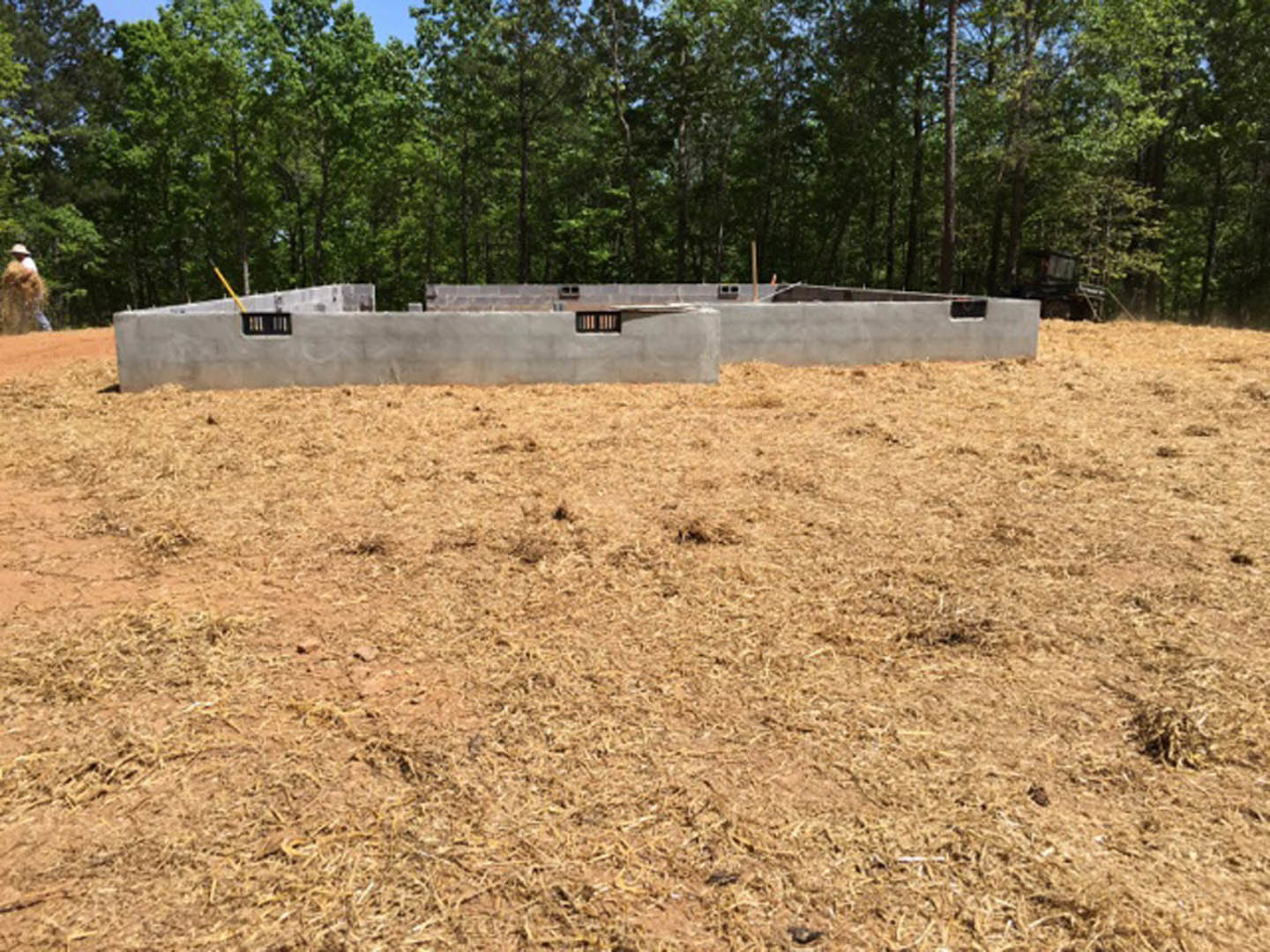 Concrete foundation set in a grassy field bordered by trees, with a partially visible concrete wall and window opening