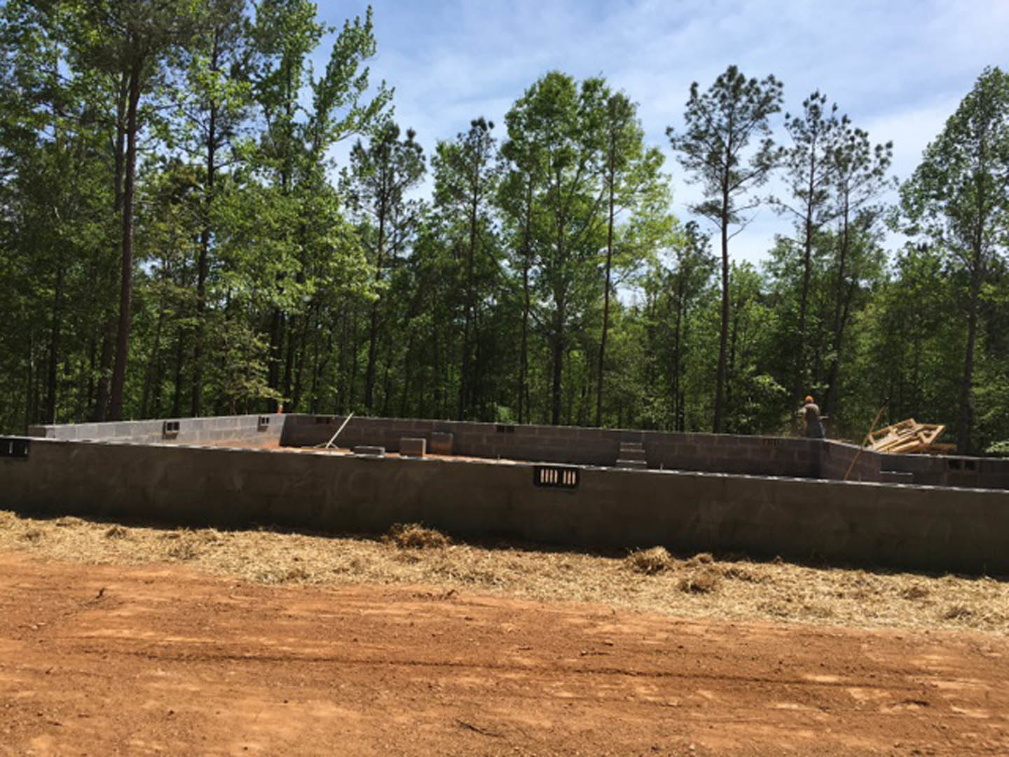 Concrete foundation slab surrounded by dirt and grass, forested area with tall leafy trees in the background, construction worker wearing a helmet standing near the edge of the
