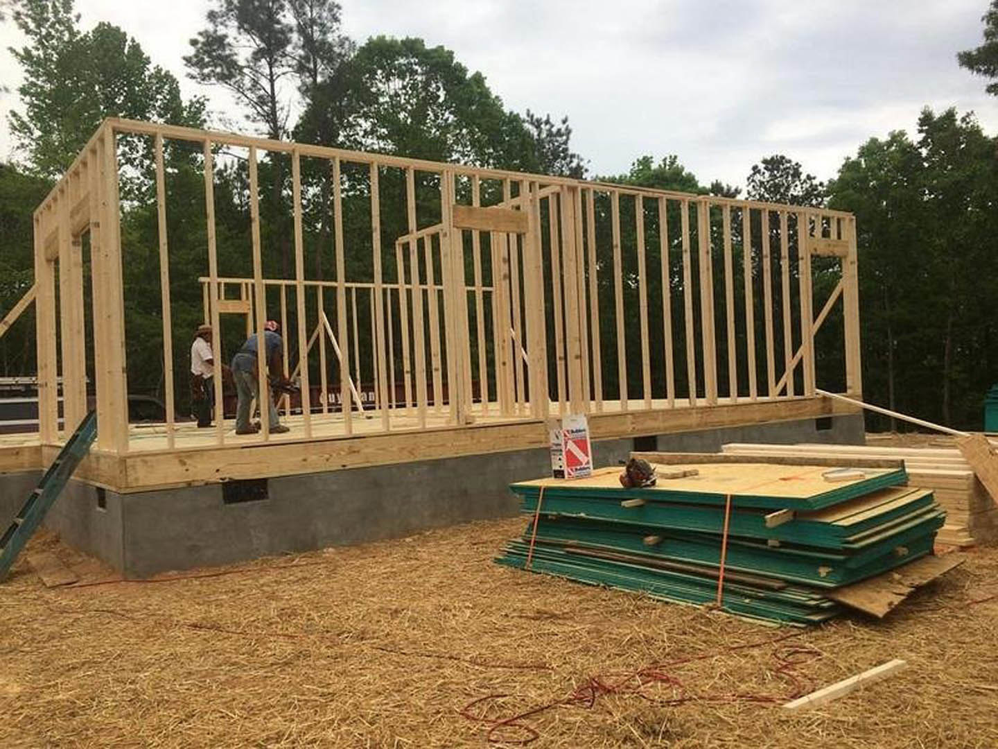 Two men assembling a wooden frame on a residential construction site, surrounded by stacked lumber and hay, with a fenced yard and trees in the background.