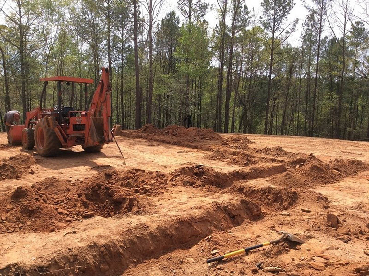 Tractor with front bucket parked on dirt construction site near wooded area, yellow and black pole in foreground, person bending over beside equipment.