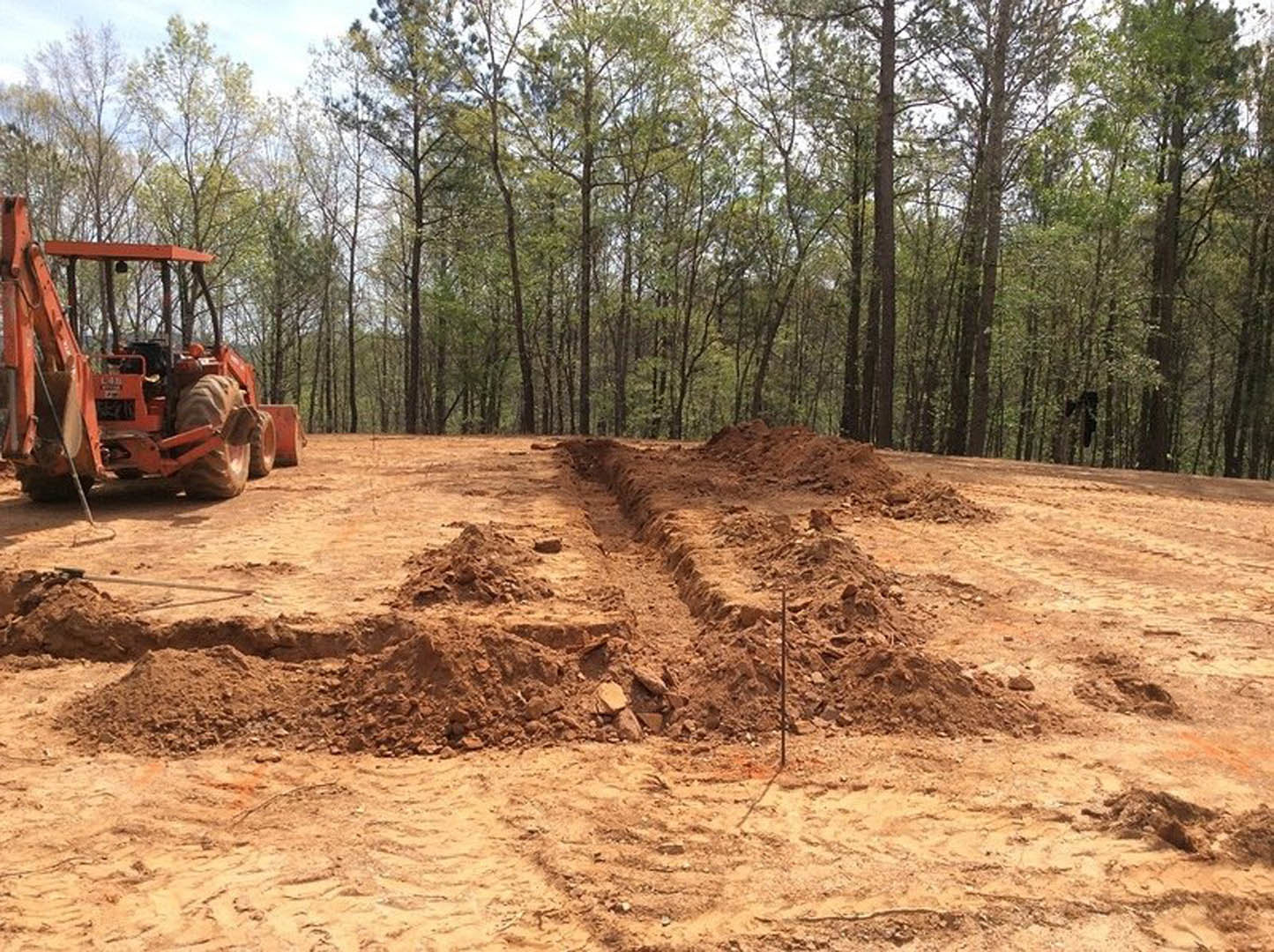 Yellow tractor parked on bare dirt lot surrounded by trees, soil tracks visible, construction equipment in foreground under blue sky