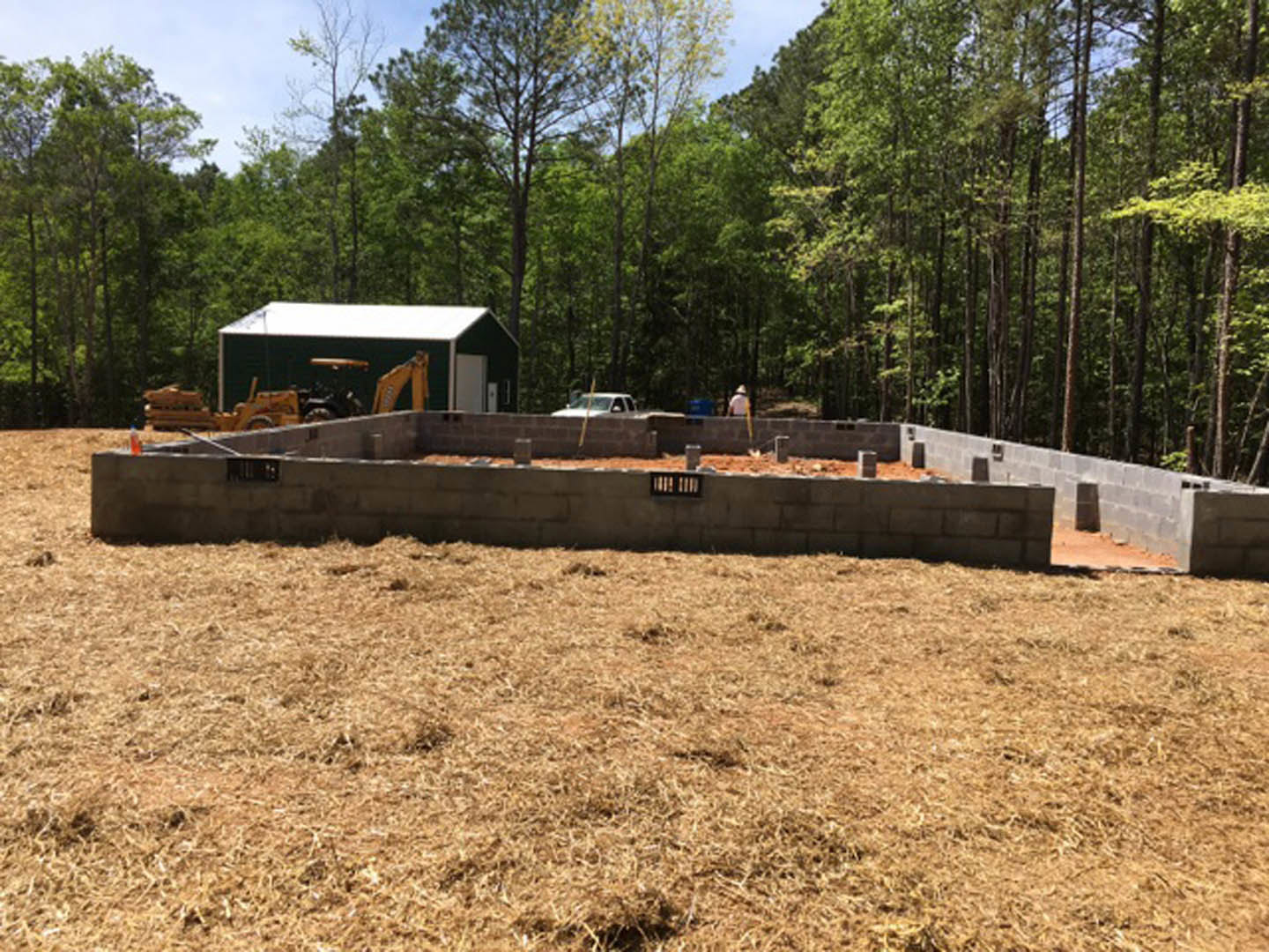 Concrete foundation wall with vent, yellow construction vehicle parked on dirt, green building partially visible among trees in background, grassy area and brick wall at edge of