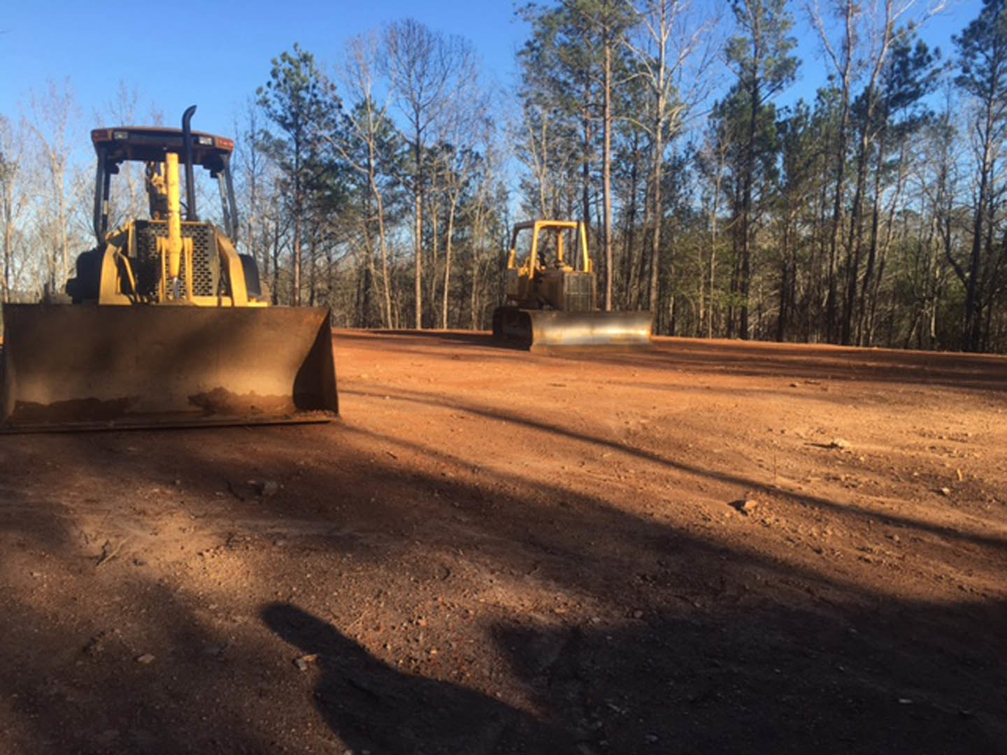 Bulldozer and construction vehicle grading dirt lot surrounded by trees under open sky