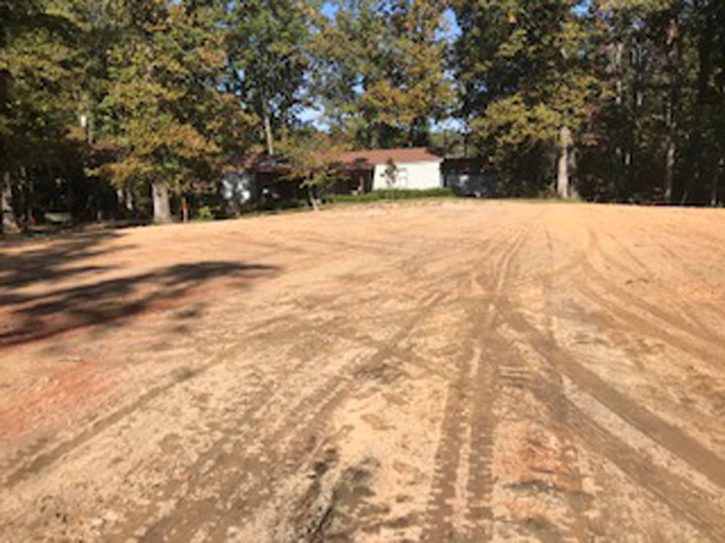 Dirt road with deep tire tracks leading toward a custom home surrounded by trees, under a clear sky