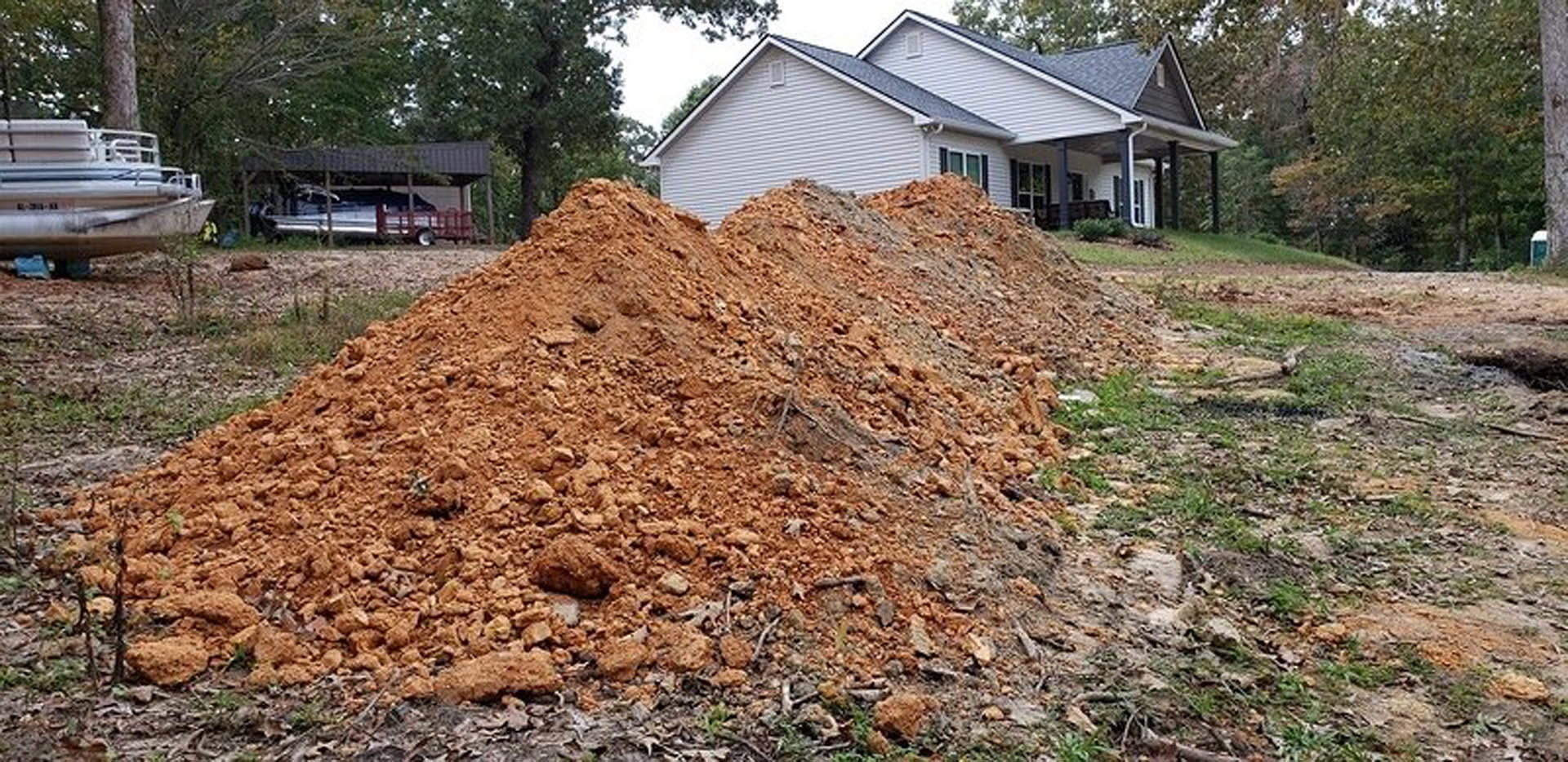 Fresh soil mound on grassy front yard, partially obscuring white custom home with gray roof, large windows, and parked car; leafy trees and blue sky in background.