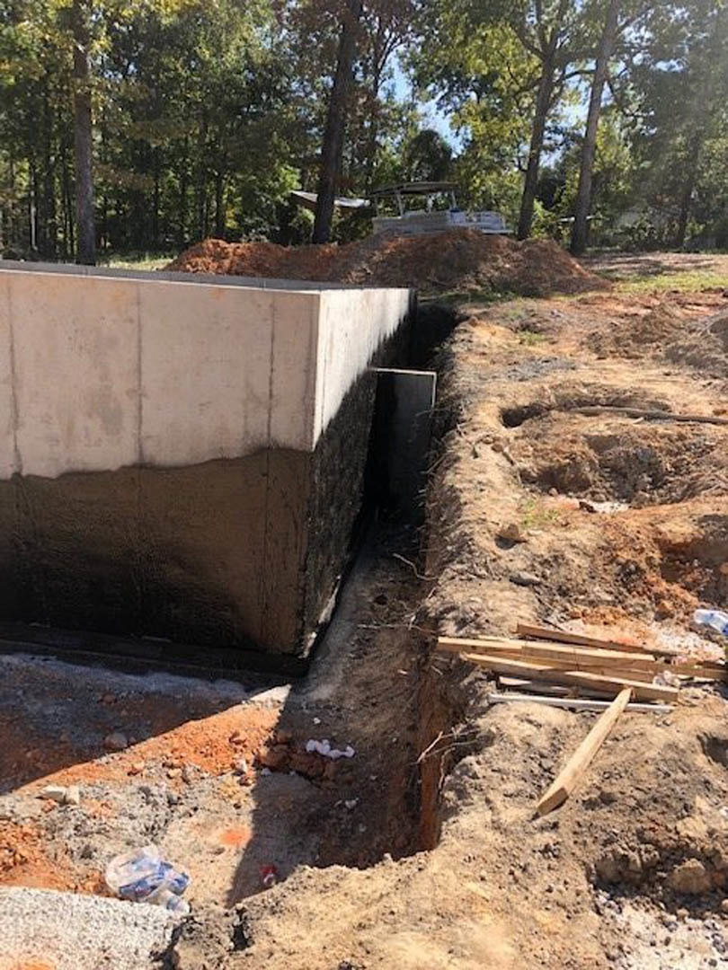 Concrete foundation wall with exposed soil and drainage hole, surrounded by dirt, rocks, and scattered wood in a wooded outdoor setting.