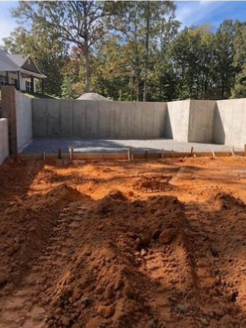 Concrete foundation and dirt mound surrounded by trees, with tire tracks on a dirt road and a person in the background.