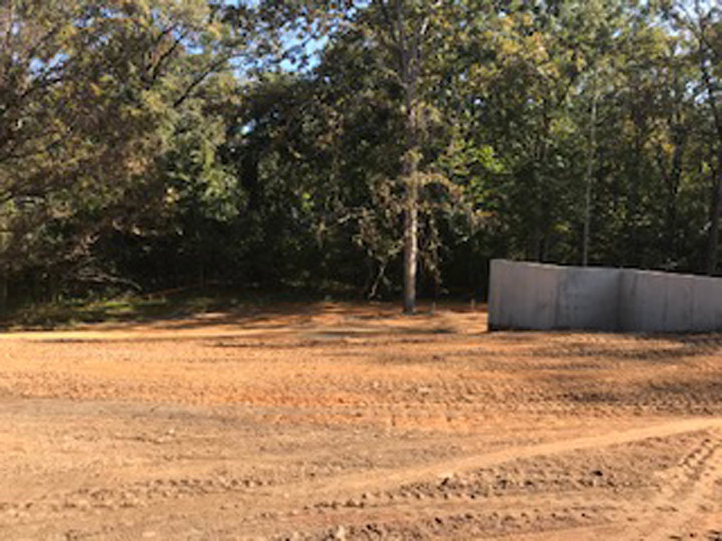 Dirt field bordered by a grey concrete wall, with mature trees in the background under an open sky
