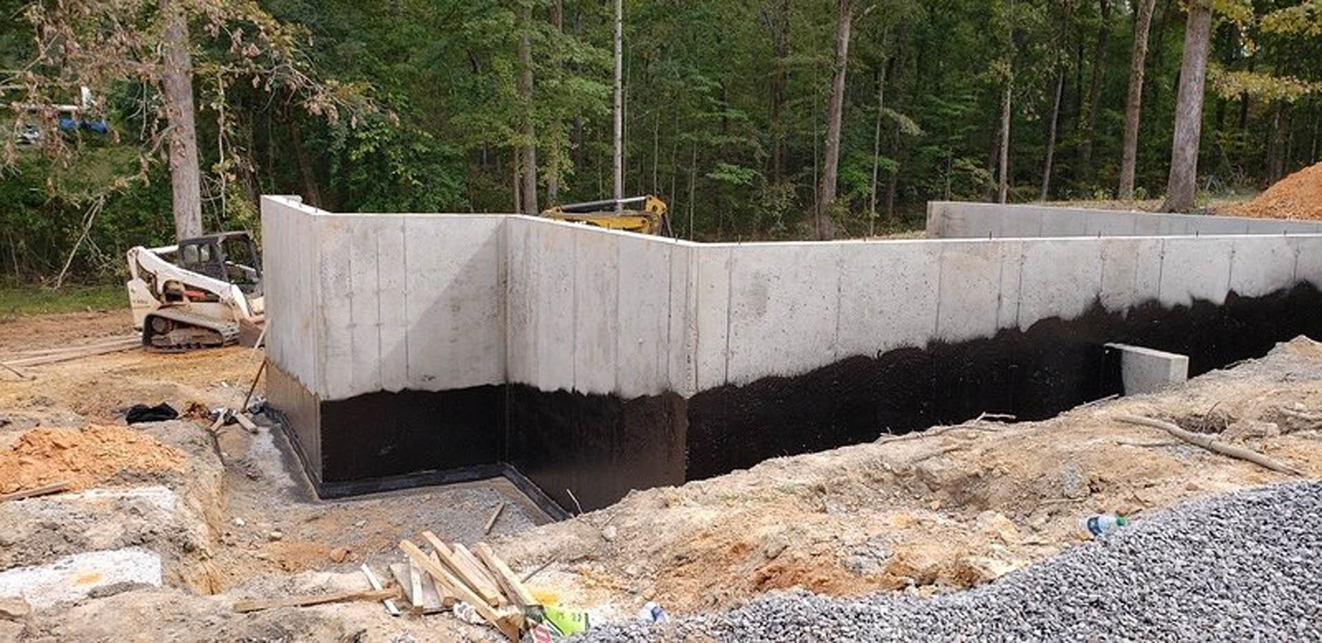 Concrete foundation with black and white painted finish, surrounded by dirt and rocks, tree and plants visible in outdoor setting, close-up of construction equipment in foreground