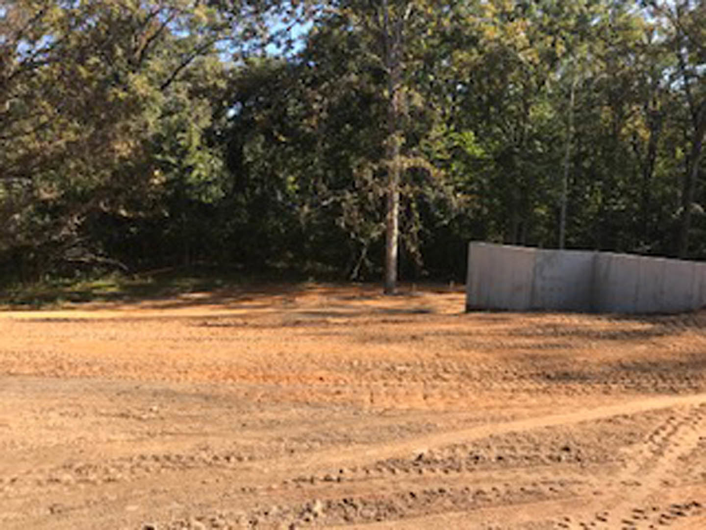 Dirt lot bordered by leafy trees, open sky above, sparse vegetation, and a person walking in the background