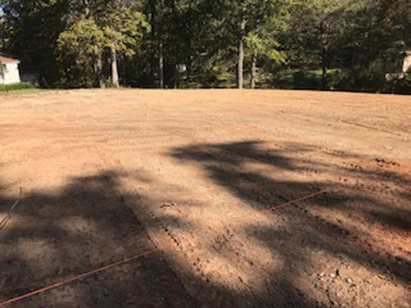 Dirt field bordered by trees, with a faint outline of a house and red marking on the ground