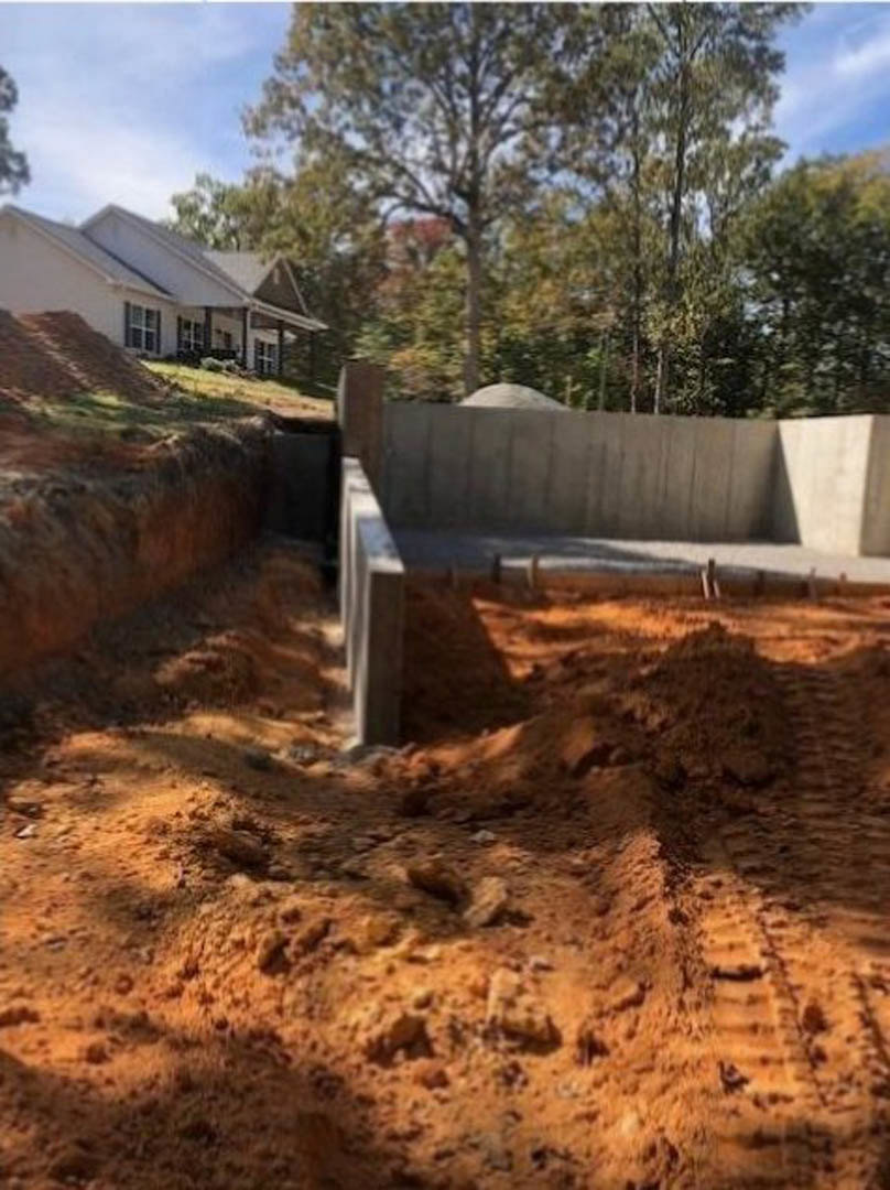 Wood-framed house under construction with exposed foundation, dirt lot, and scattered building materials; person standing near temporary fence; cloudy sky and trees in background