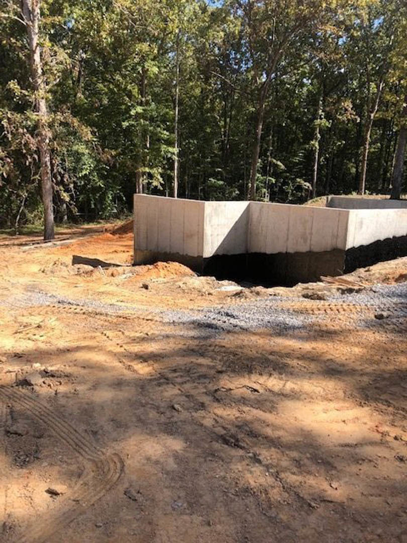 Concrete foundation with black waterproofing on exterior wall, surrounded by dirt area with tire tracks and trees in background