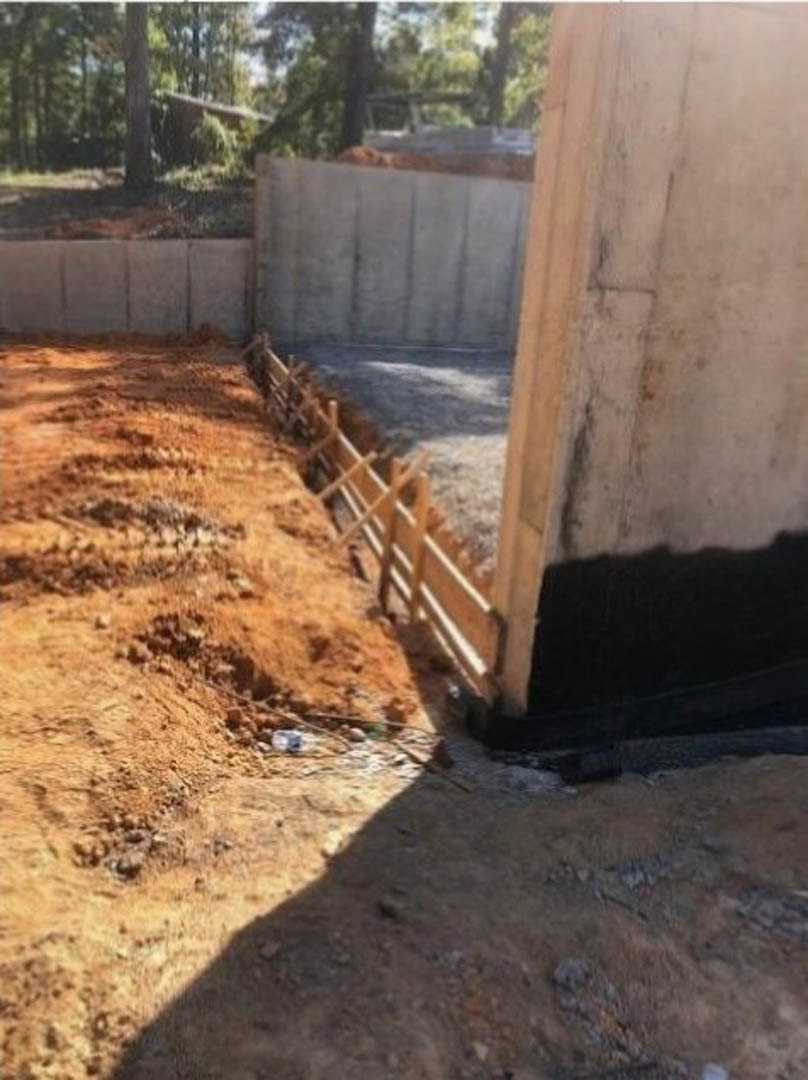 Wooden construction fence bordering dirt and rocks, foundation work visible, trees and river in background