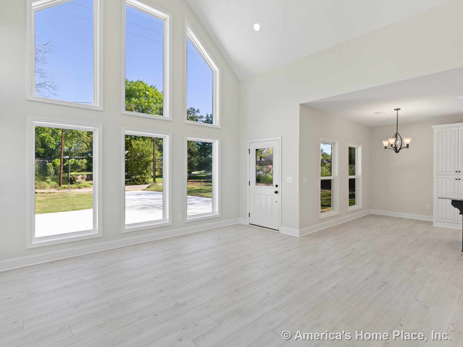Vaulted ceiling living area with a wall of tall windows, light wood flooring, white trim, glass-paneled entry door, built-in cabinetry, and open layout connecting to dining space.