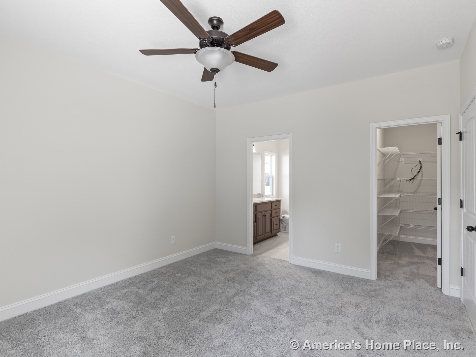 Bedroom with gray carpet flooring, ceiling fan with light, direct access to walk-in closet featuring wire shelving, and bathroom with wood cabinetry vanity; white trim and doors