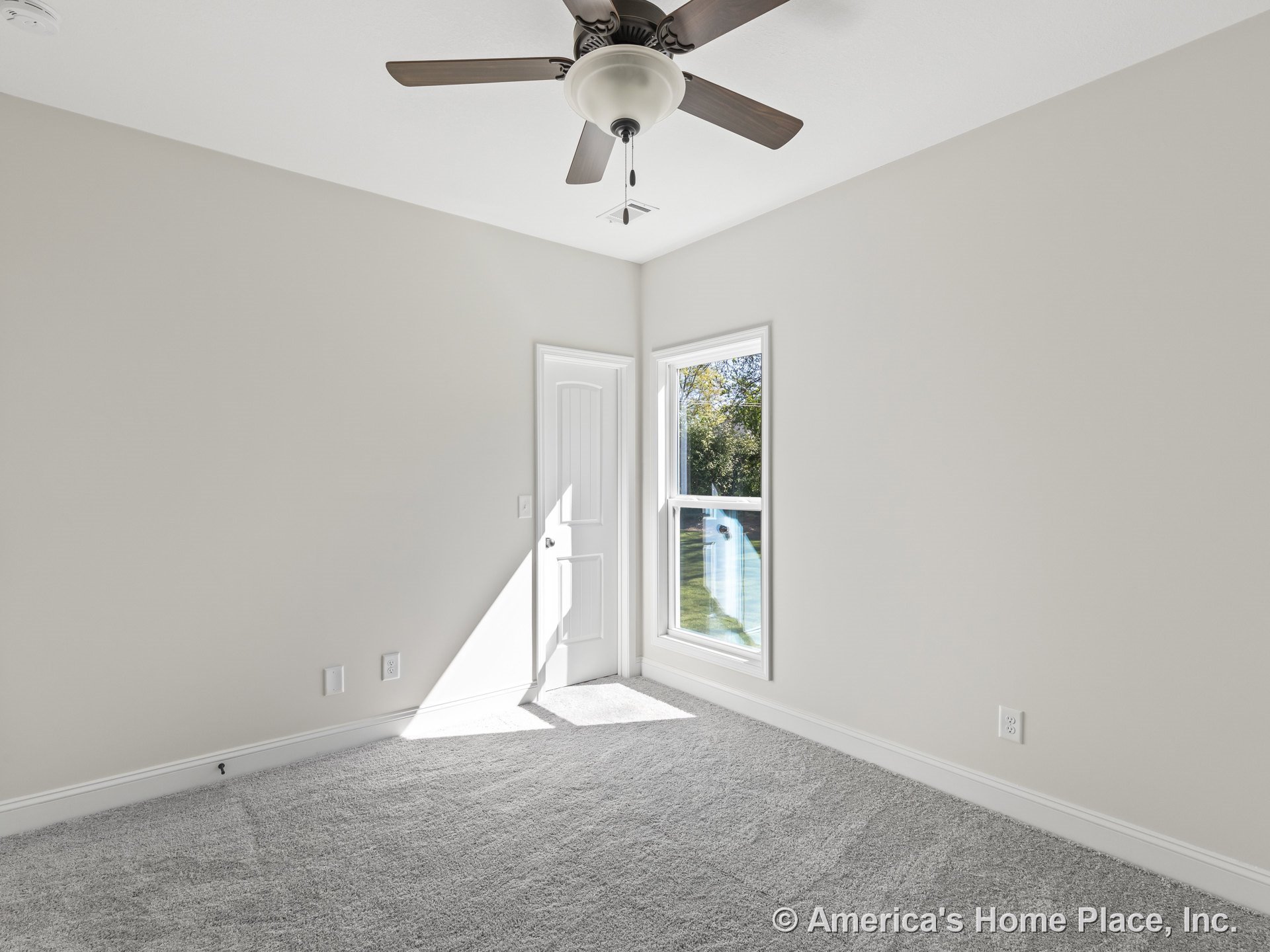 Bedroom with light gray carpet, neutral painted walls, white baseboard trim, ceiling fan and light fixture, large double-hung window allowing natural light, and white paneled door.