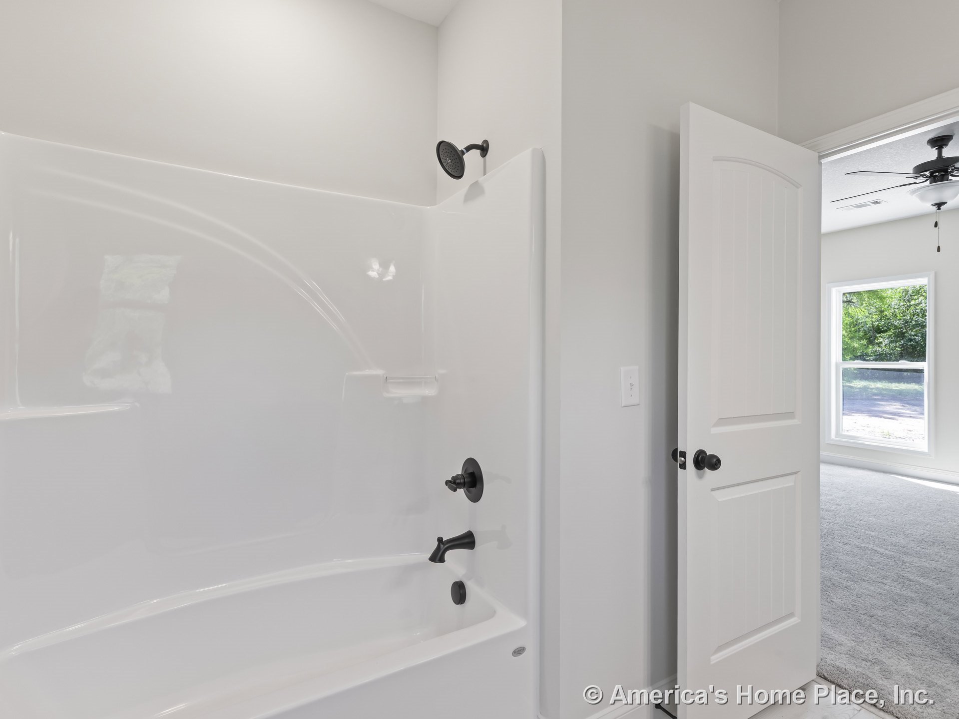 White fiberglass tub and shower with matte black showerhead and faucet, white paneled door leading to adjacent room with gray carpet, large window with white trim, neutral walls.