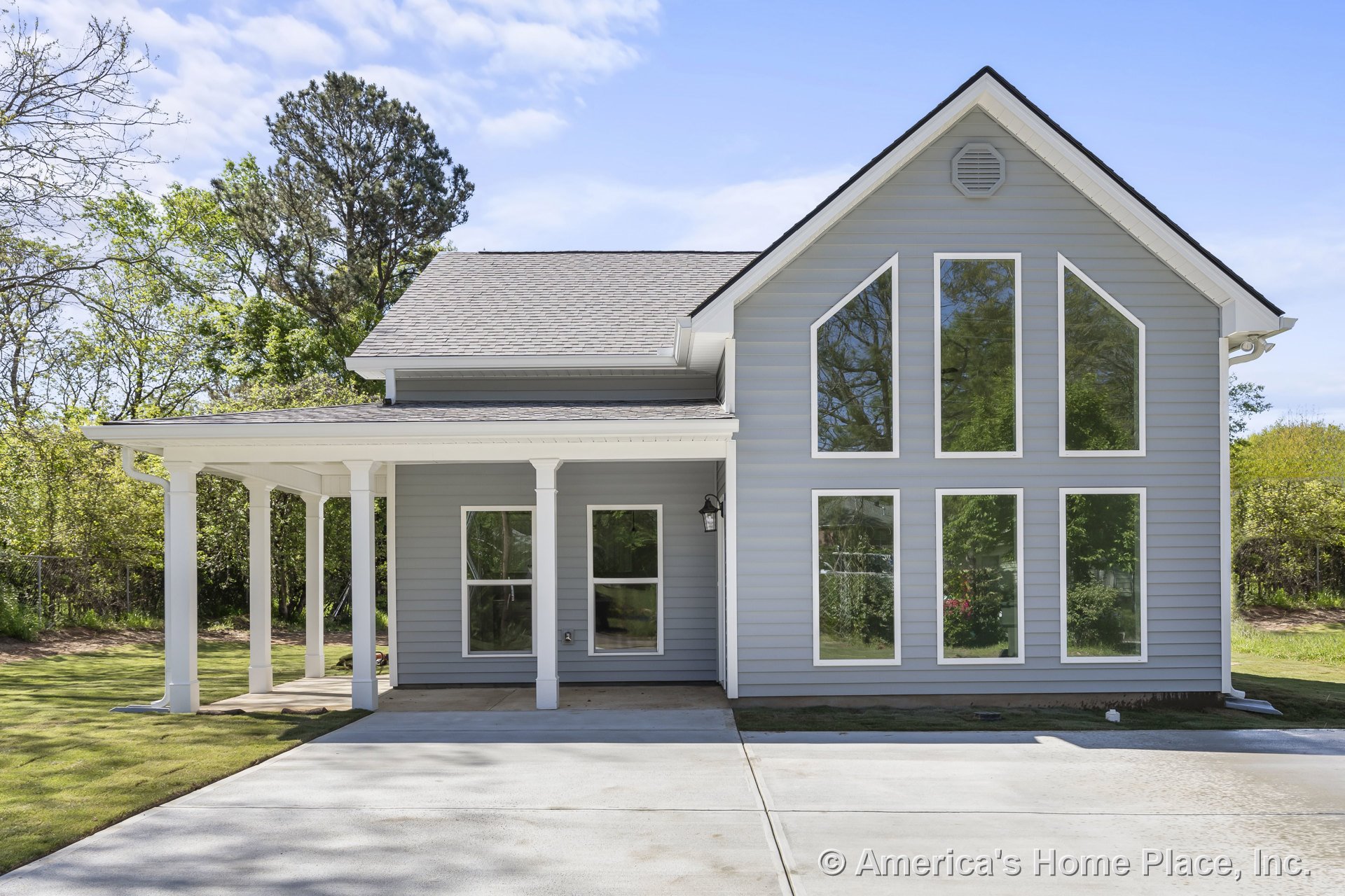 Gray horizontal siding with prominent geometric windows, covered front porch supported by white columns, concrete driveway, gable roof with white trim and permanent lighting.
