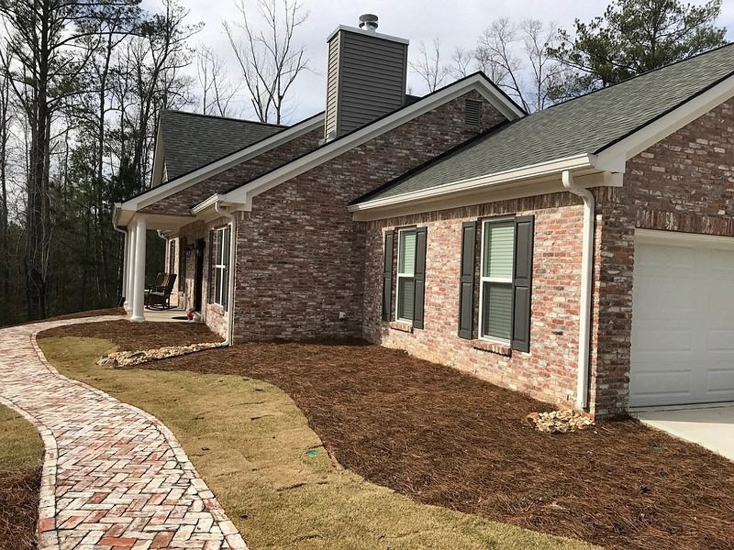 Red brick house with white-framed windows, white brick accent wall bordered in black, brick walkway bordered by grass, outdoor chair near entrance, window with closed blinds
