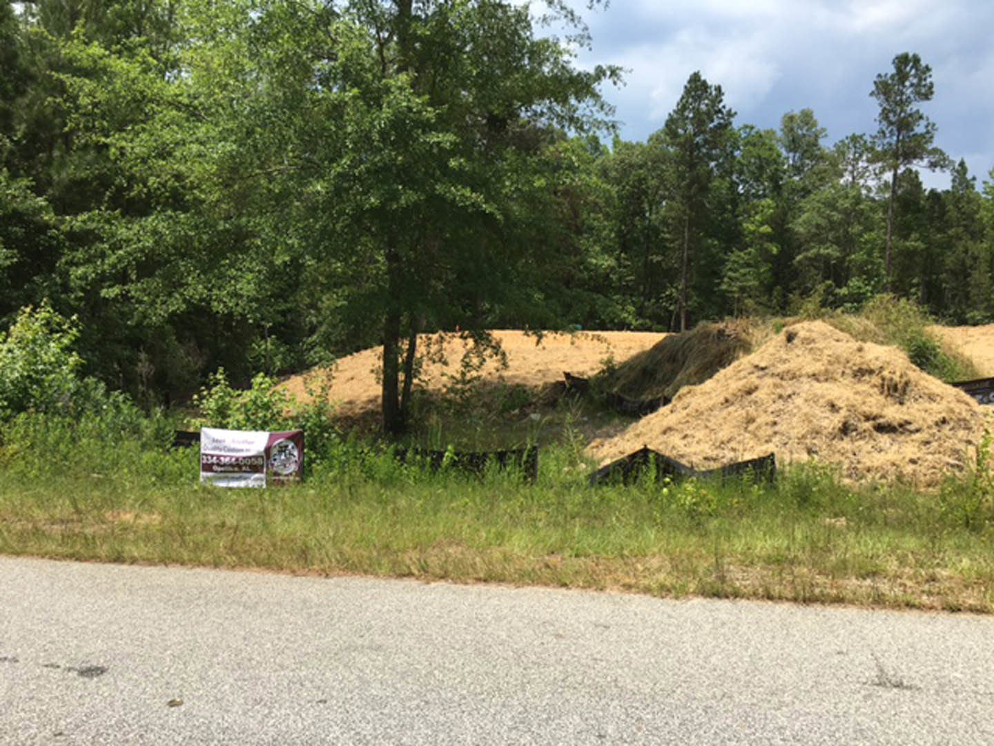 White roadside sign surrounded by grassy field, dirt hill, and cluster of trees beneath blue sky with scattered clouds