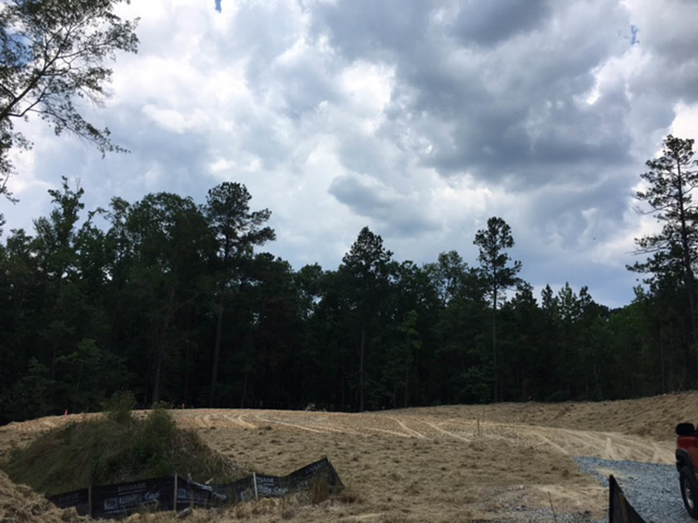 Dirt field bordered by a wooden fence, scattered grass patches, and dense trees under a cloudy sky