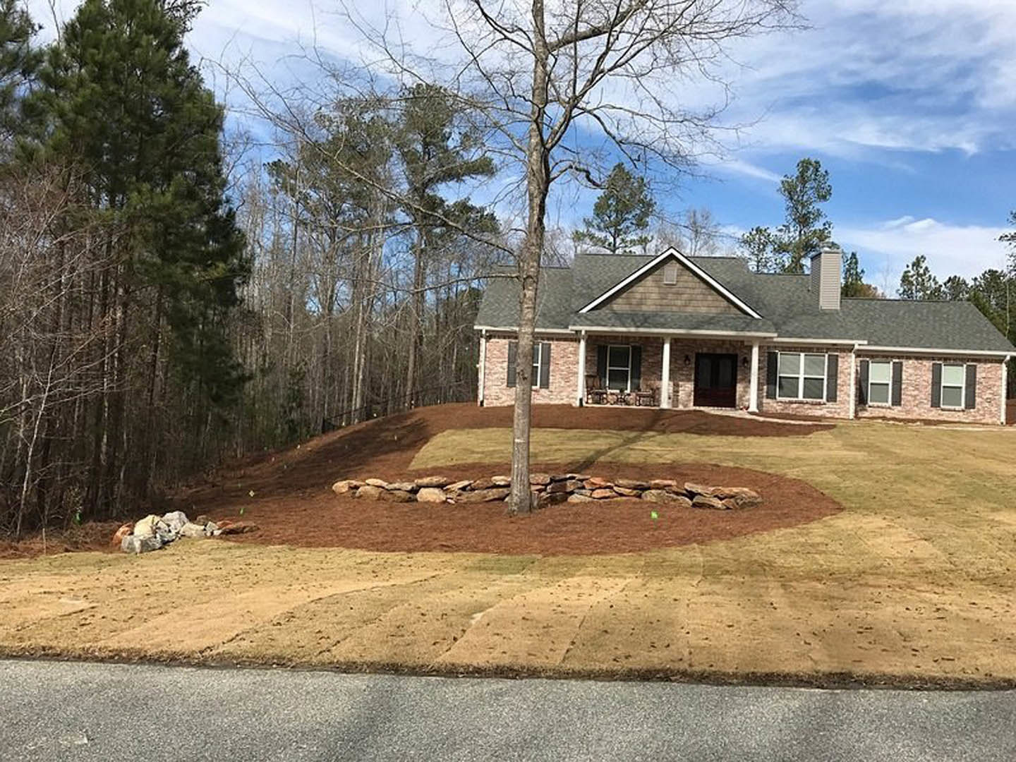 Two-story house with white-framed windows, covered front porch, brick chimney, and mature tree surrounded by a stone retaining wall in landscaped yard