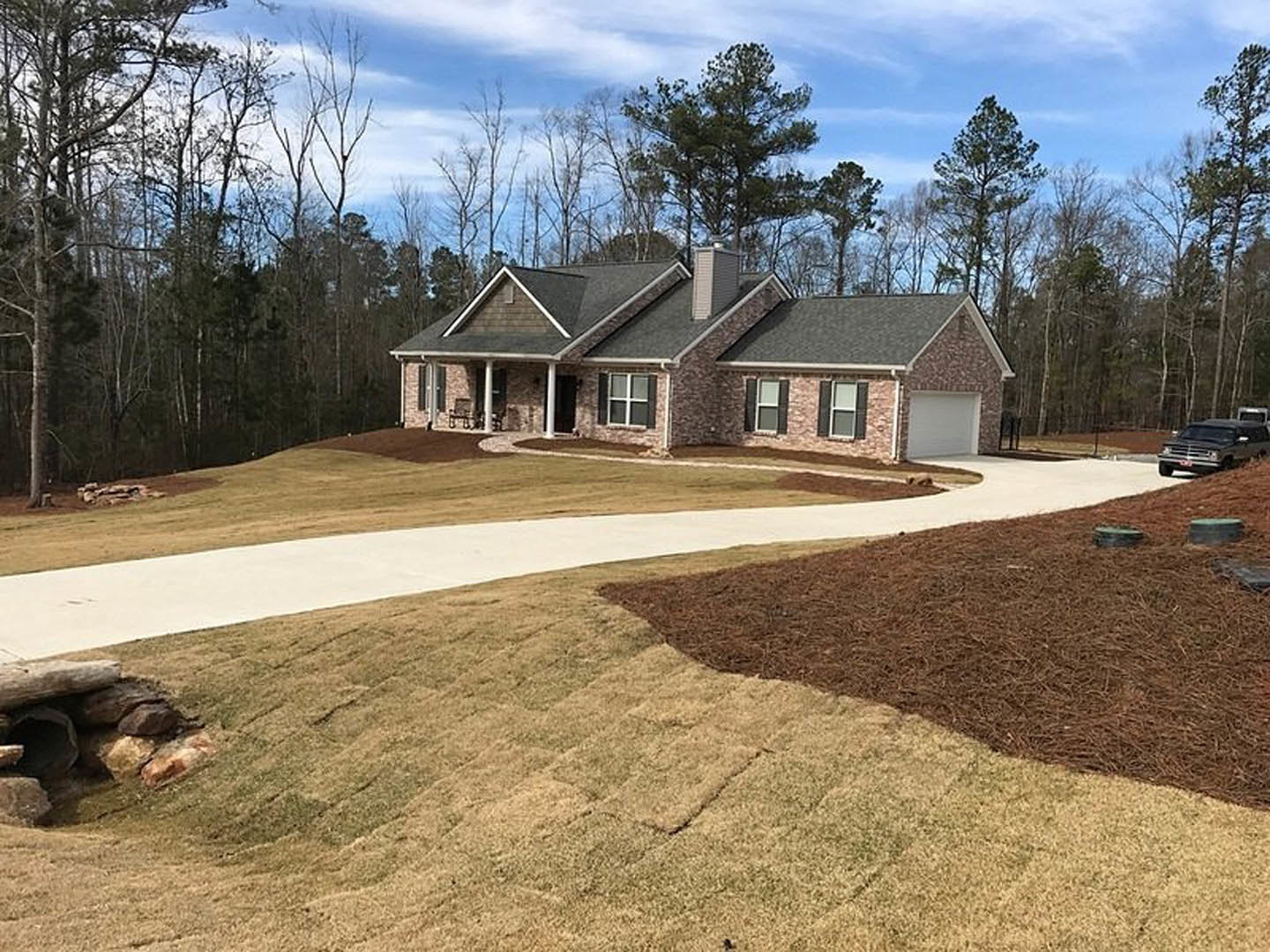 White house with black roof, brick chimney, paved driveway, mature trees, brown grass and mulch, rocks and logs in front yard, car parked roadside