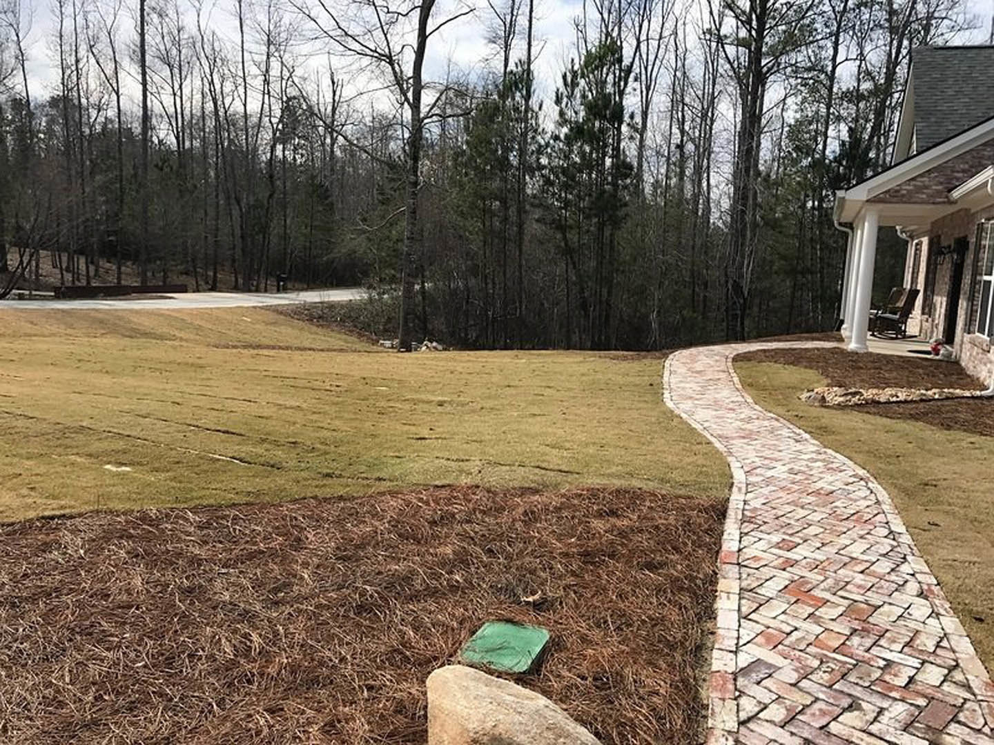 Brick walkway bordered by grass and dirt alongside a house with landscaping and lawn visible