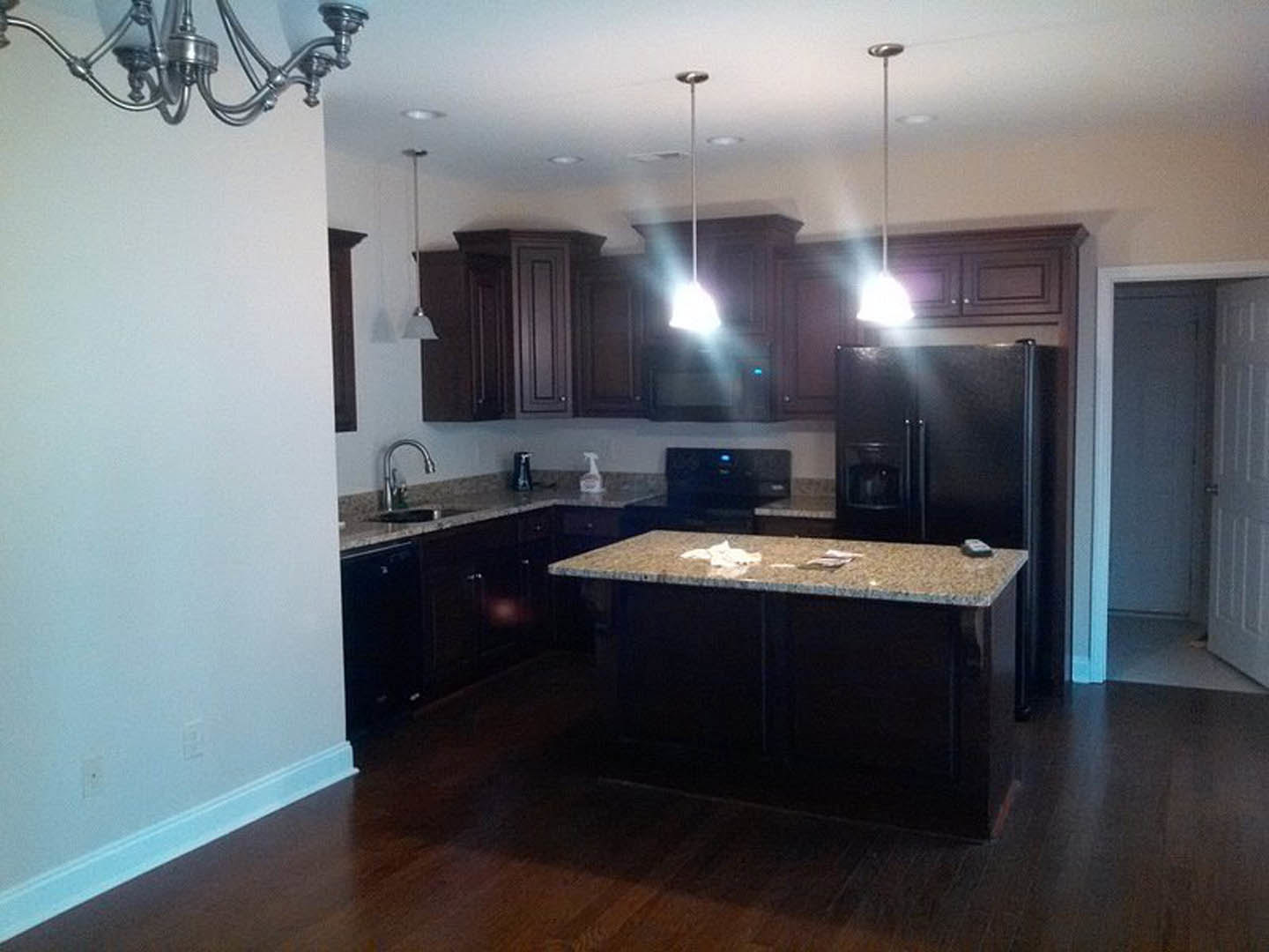 Kitchen with dark wood cabinets, granite island countertop, stainless steel sink, pendant chandelier, light-colored tile flooring, papers and phone on the island, neutral painted