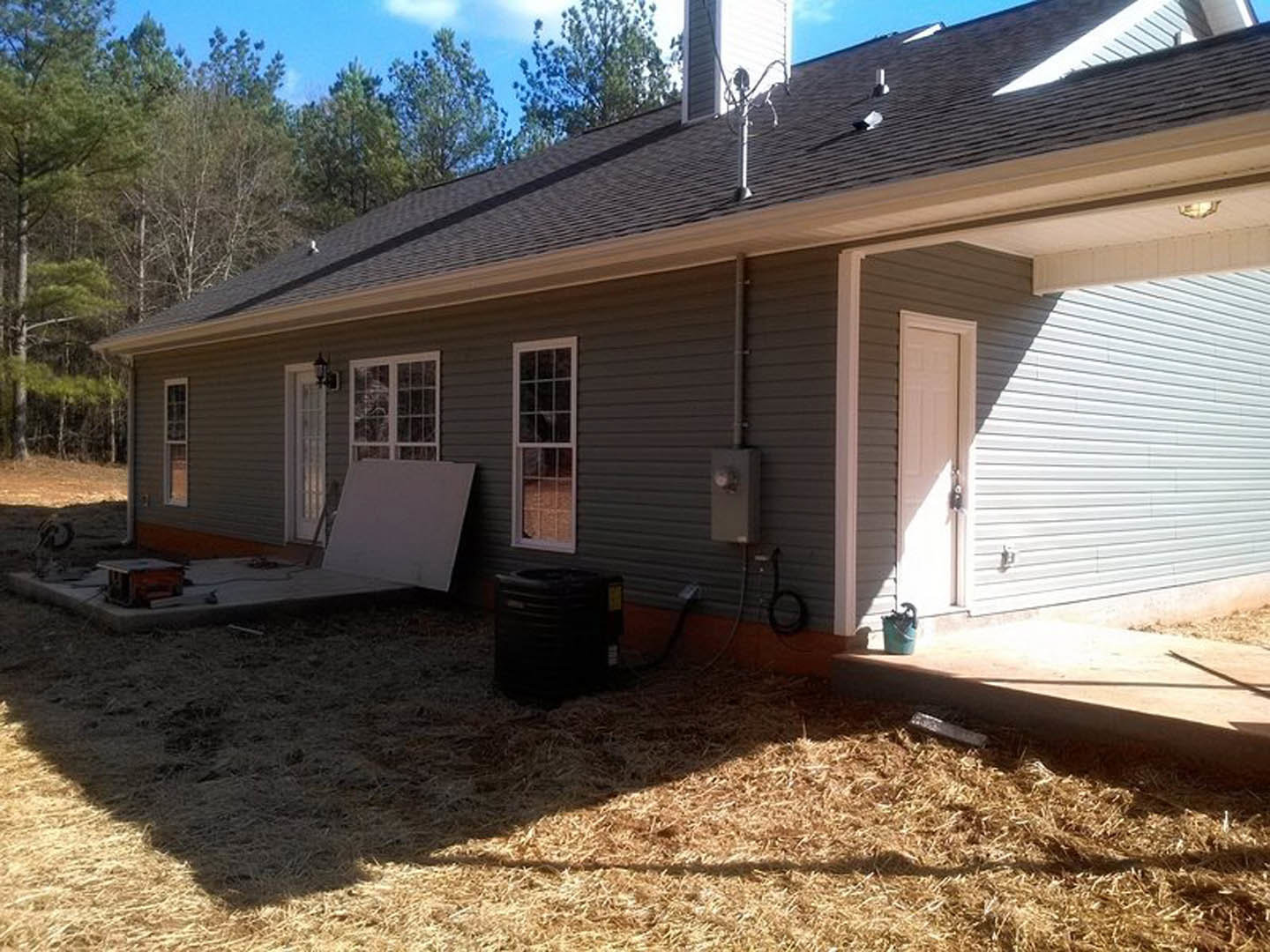 White-paneled house with open white door illuminated by porch light, multi-pane window, black trash can on ground, tree and blue umbrella visible in yard