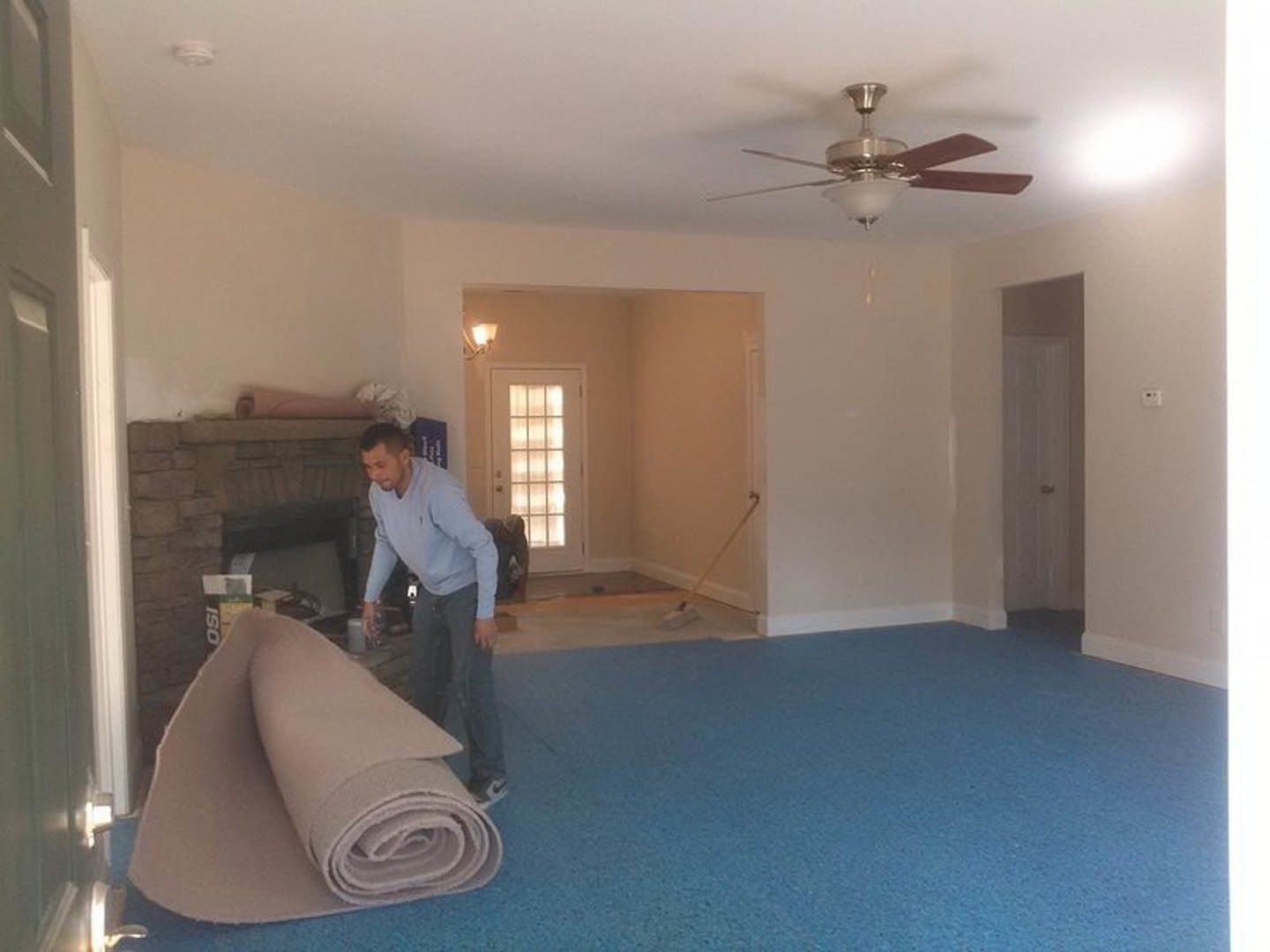 Man in blue shirt standing on beige carpeted floor, ceiling fan with light fixture above, white door with window, wall-mounted light fixture, blue accent surface, plaster walls.