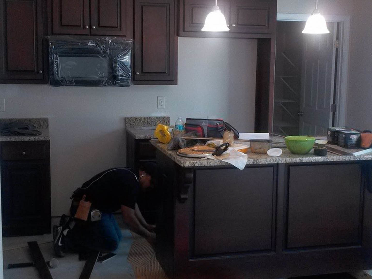 Man kneeling beside white kitchen cabinetry, plastic-wrapped microwave on countertop, green bowl with lid, plate of food, blurry bag in foreground