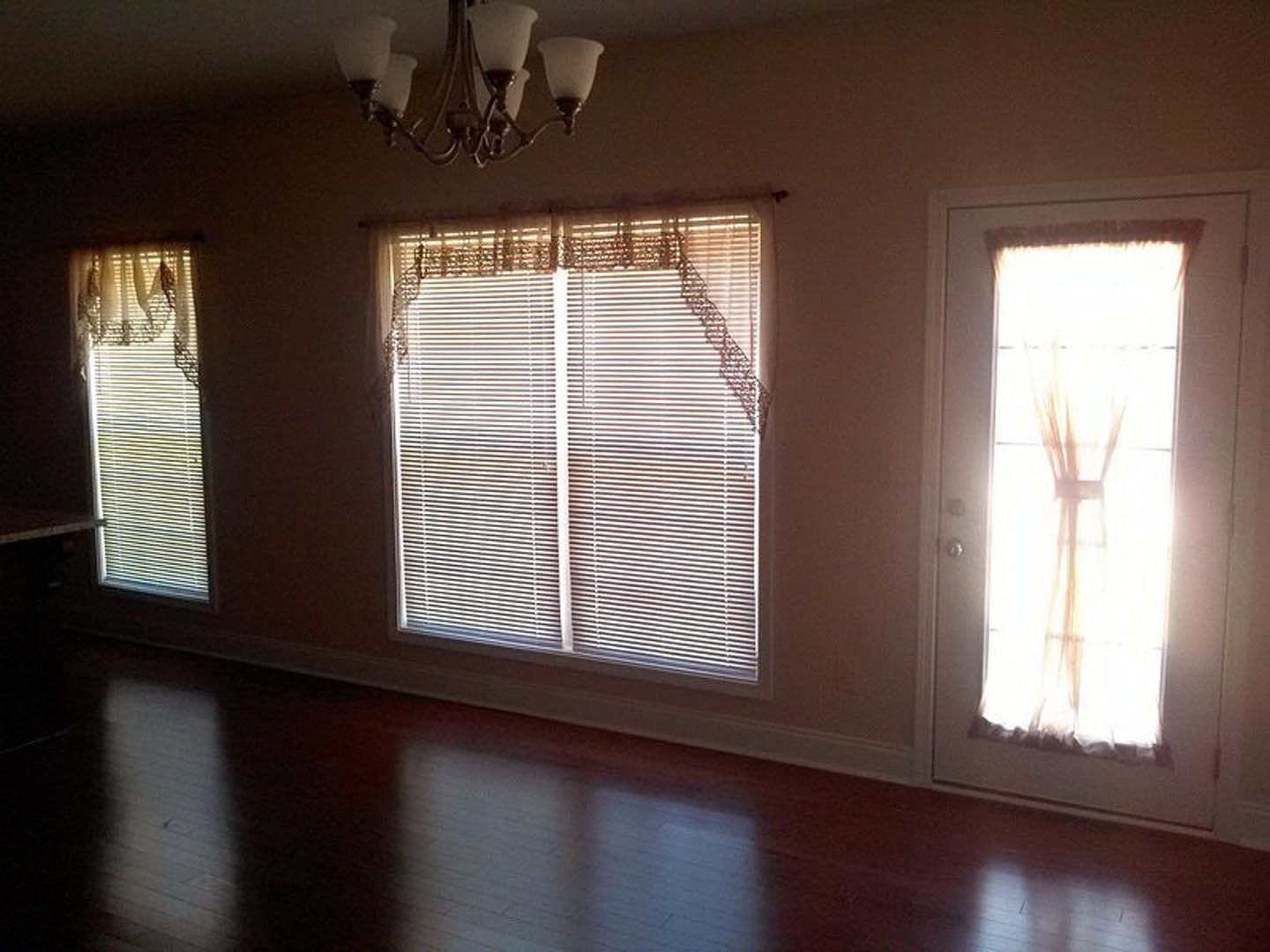 Neutral-toned room featuring a door with a curtain, two windows with white blinds, light wood flooring, and a close-up of a modern chandelier.