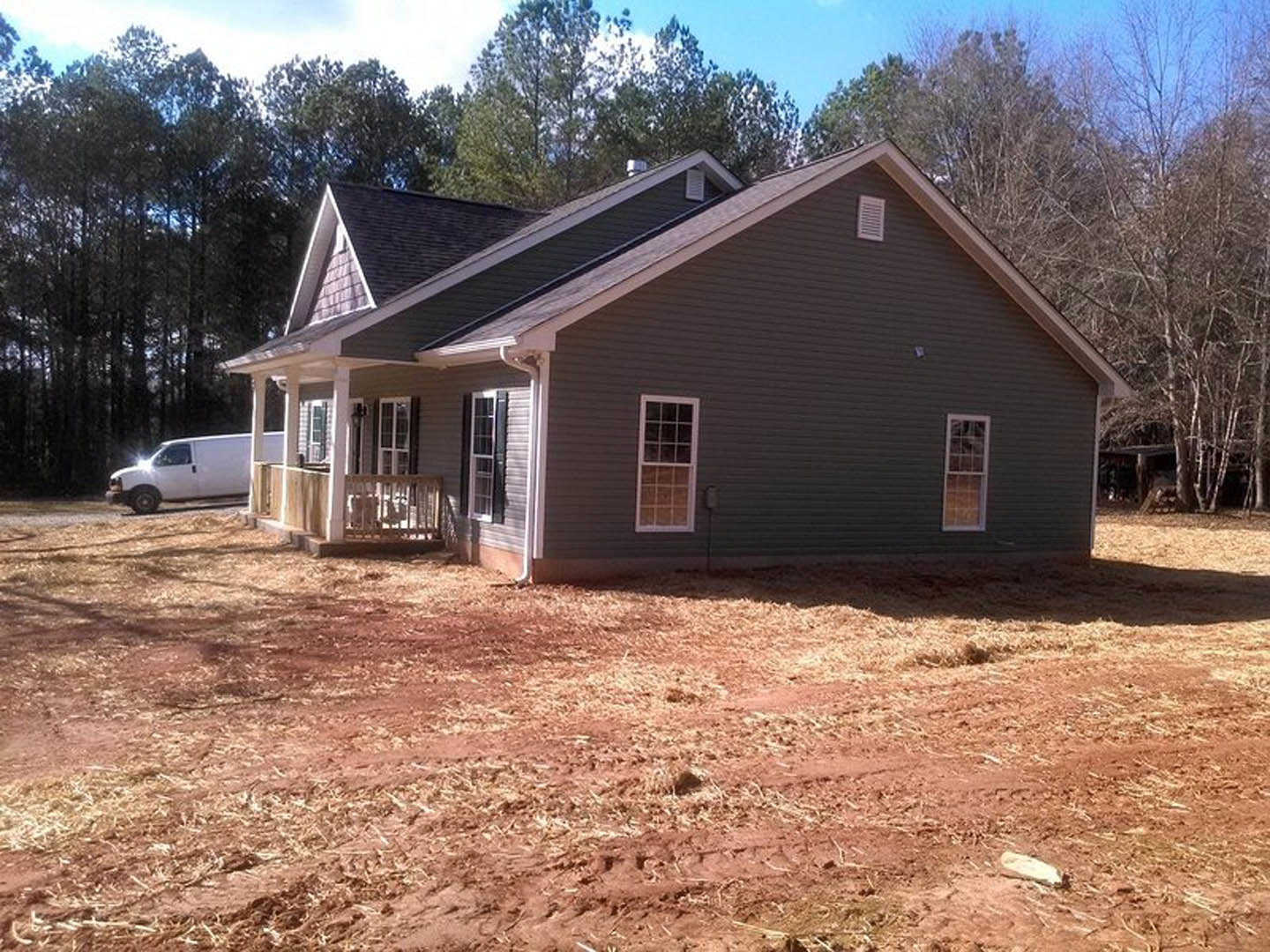 Two-story house with gray siding, covered front porch, white framed windows, metal railing, patchy grass yard, and a white van parked behind the home.
