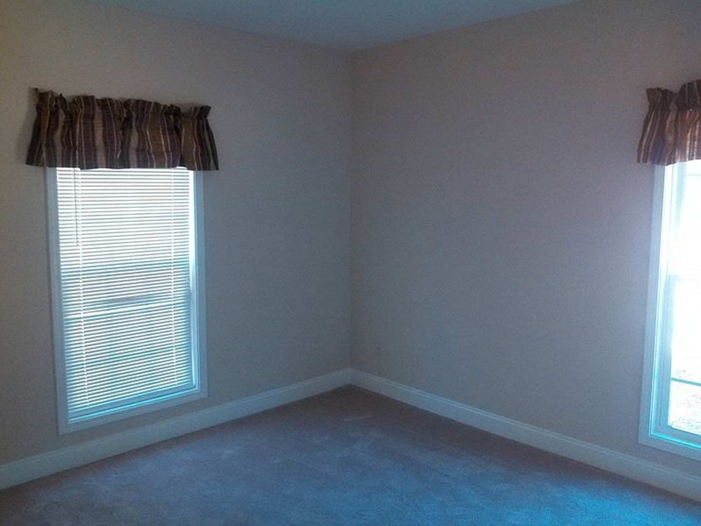 Sunlit room featuring a large window with white blinds, striped pillow resting on the windowsill, grey flooring, and neutral plaster walls.