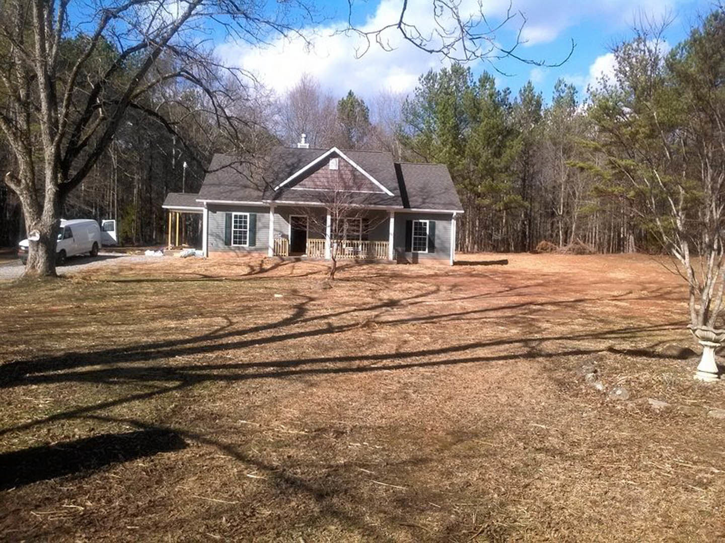 Two-story house with wide front porch, expansive grassy yard, mature trees in background, white van parked beside tree, multiple square windows, cloudy sky overhead