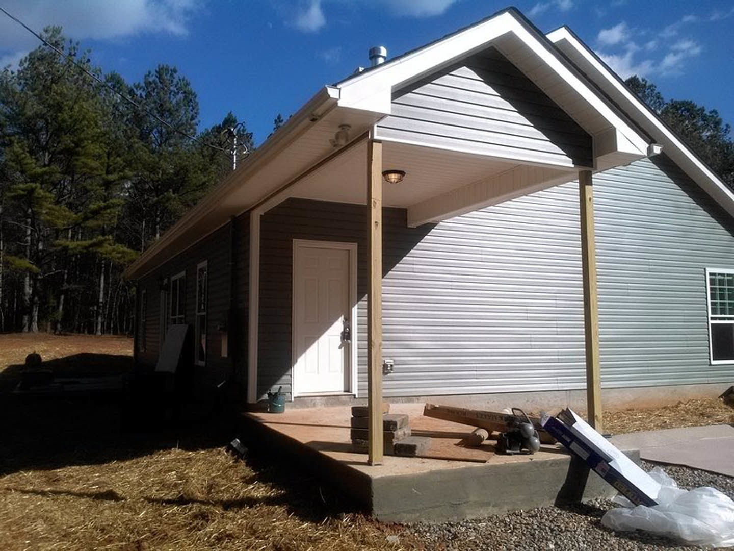 Two-story home with light siding, covered front porch, white framed windows, white entry door with handle, book resting on porch ledge, plastic bag on ground, tree and cloudy sky
