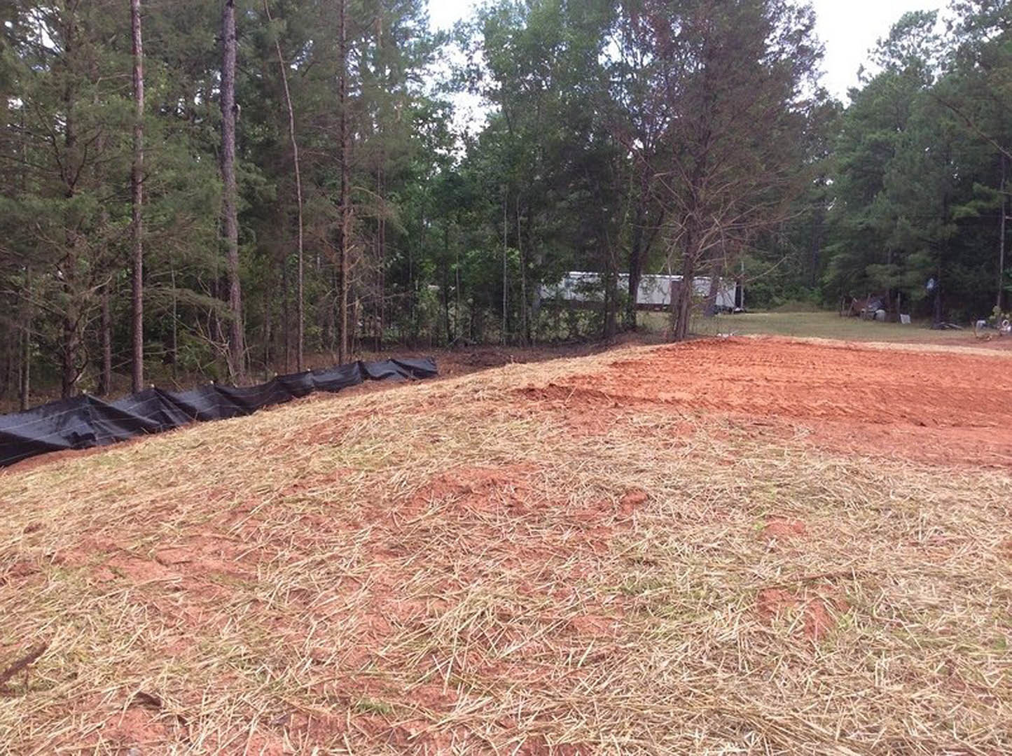 Dirt field scattered with black plastic bags, surrounded by grass and trees under a partly cloudy sky
