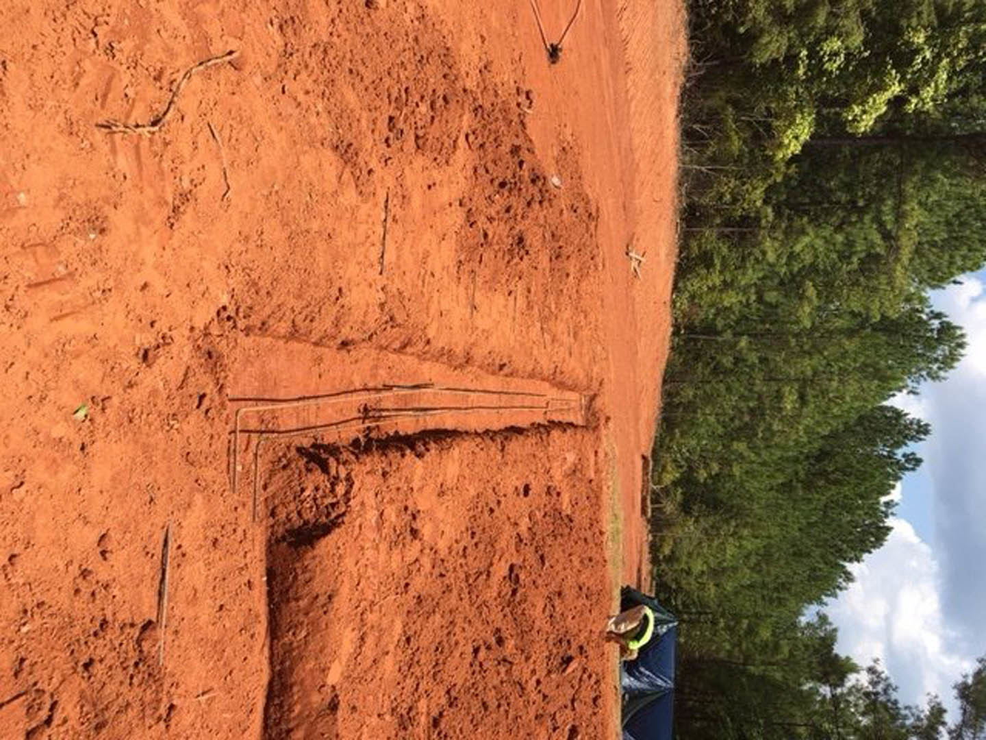 Dirt field bordered by tall trees under blue sky with scattered clouds