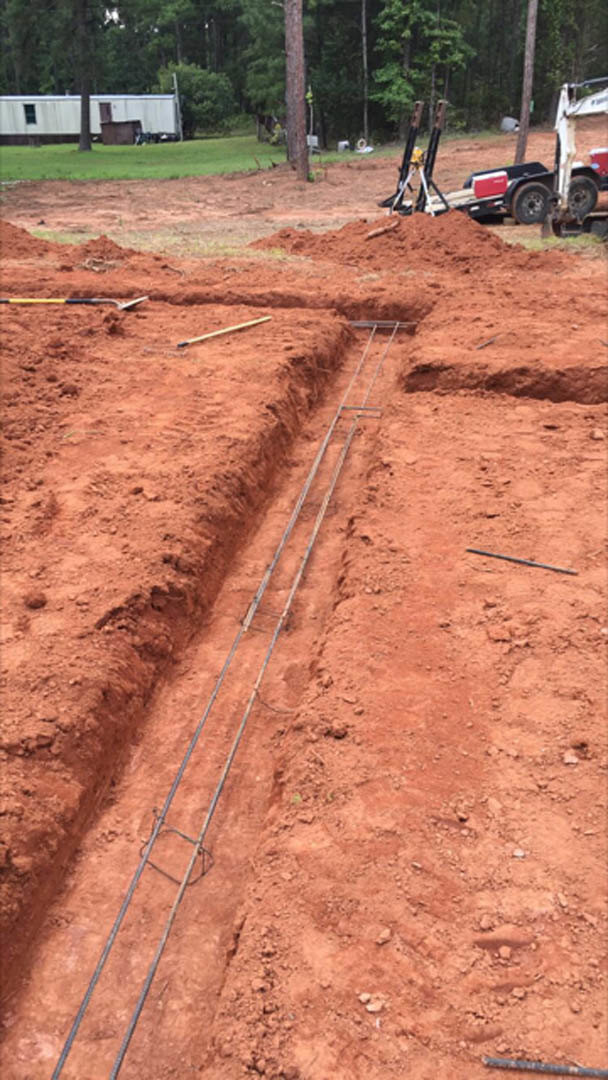 Wide dirt trench running through a muddy yard with tire tracks, white trailer and crane in the background, trees lining the edge of the property.