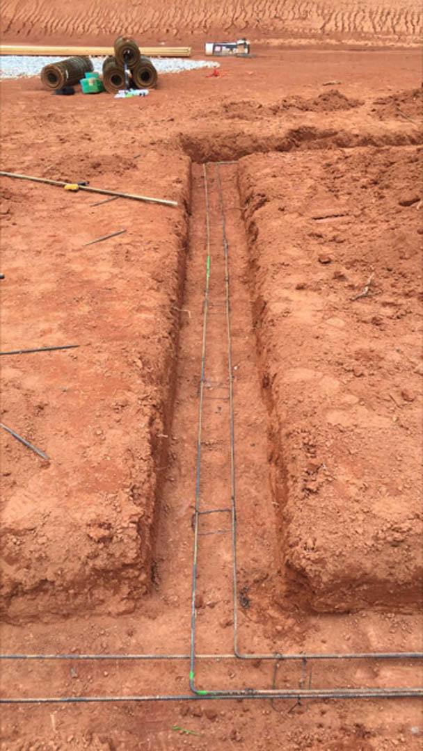 Freshly dug trench in brown soil with a hammer resting nearby, rolls of material visible along the edge, outdoor construction site adjacent to brick exterior.